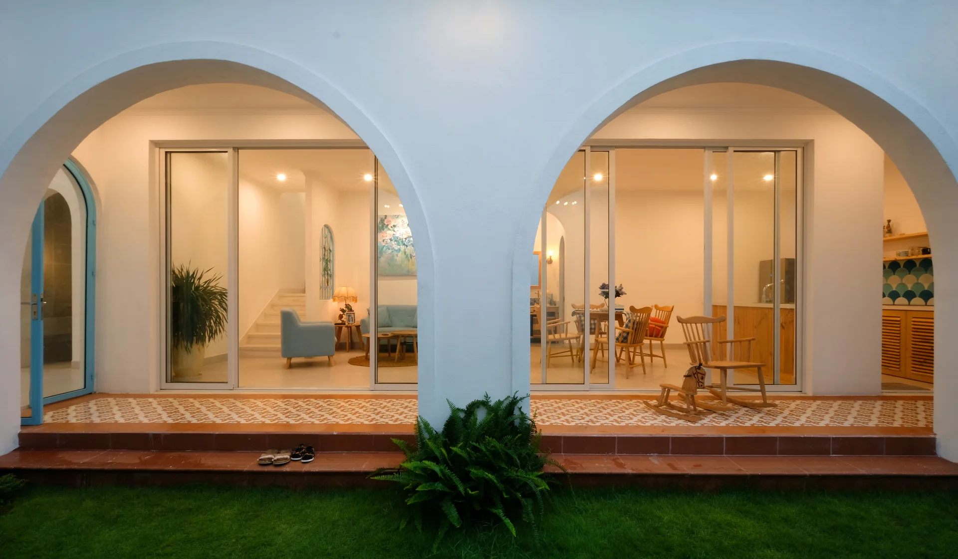 A white arched portico frames large glass sliding doors revealing a brightly lit home interior, with geometric patterned tiles on the patio floor.