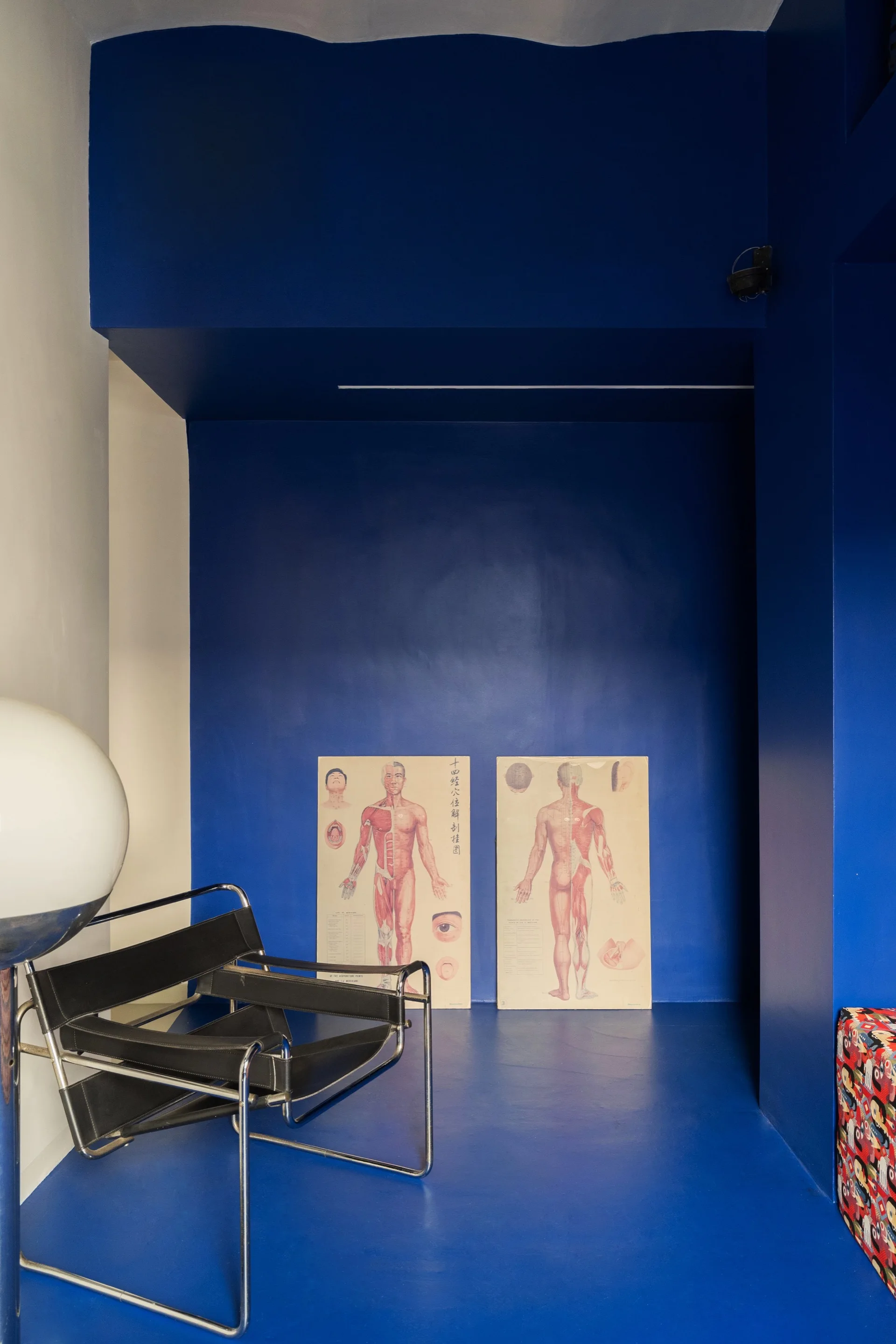A deep blue painted alcove features a black leather and chrome lounge chair, a globe floor lamp, and two anatomical chart posters leaning against the wall.