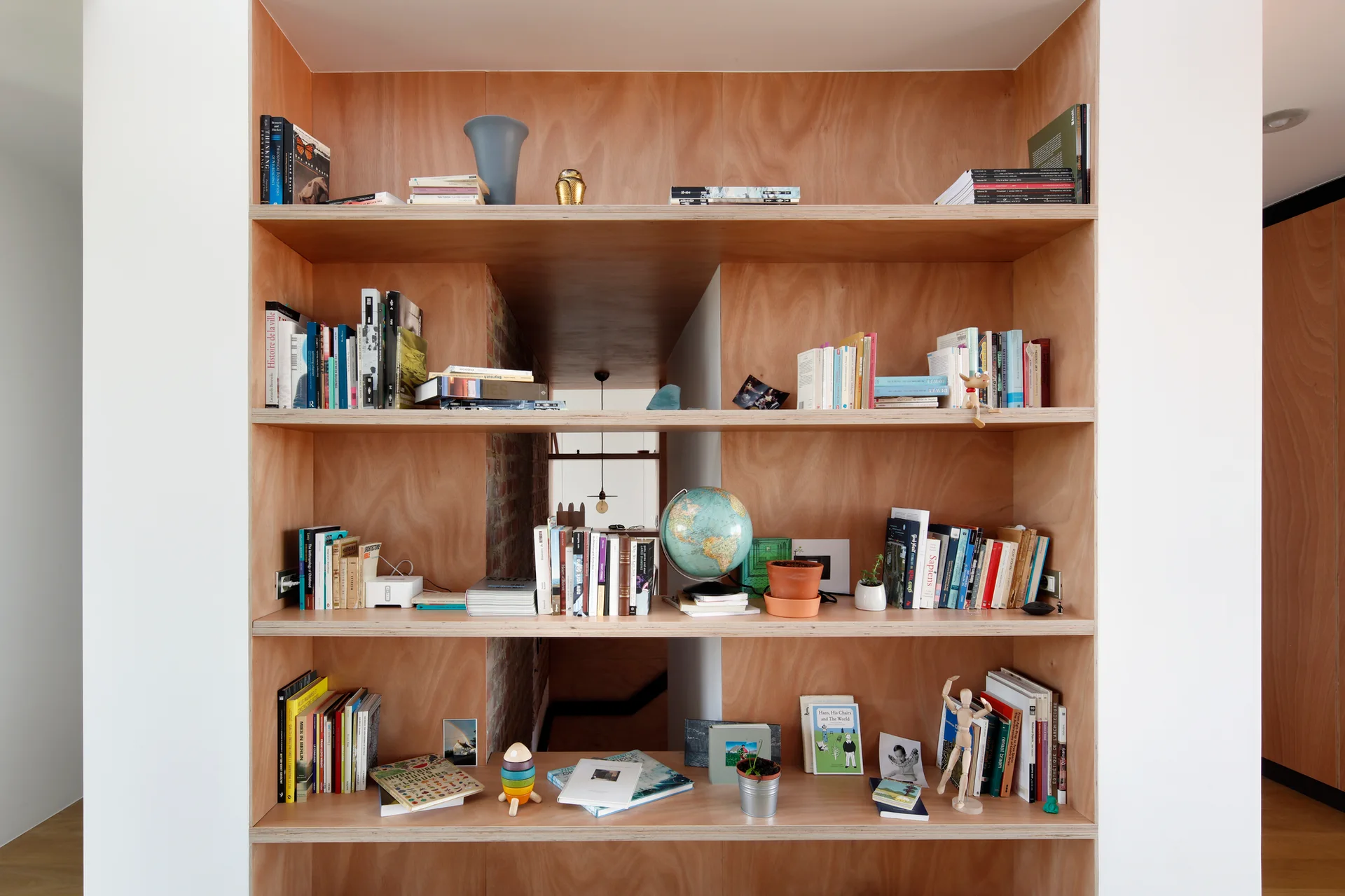 A built-in plywood shelving unit filled with books and small decor features open cutouts showing an exposed brick wall behind it.