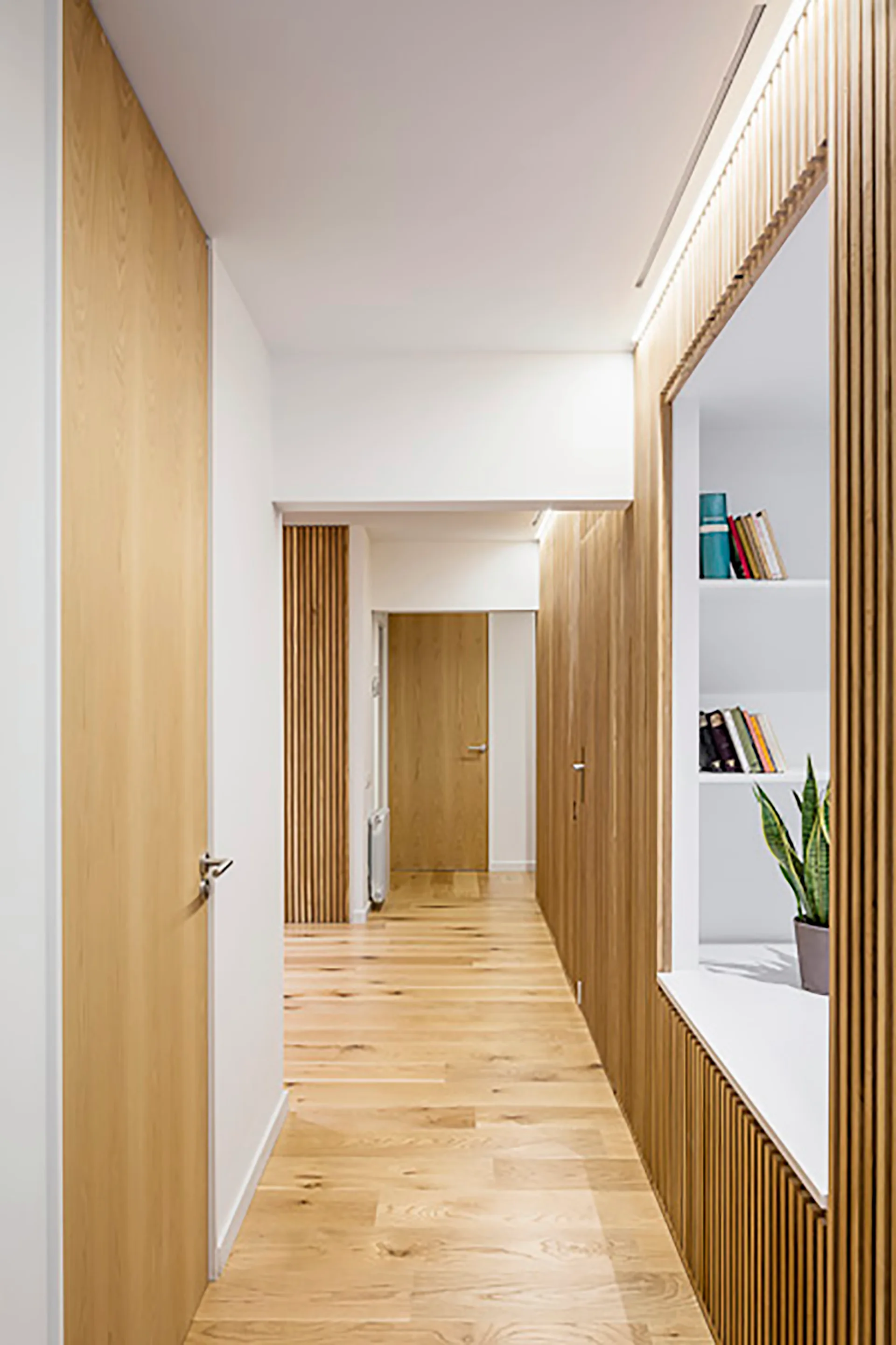 A minimalist hallway features light wood plank flooring, vertical wood slat paneling, a white recessed bookshelf, and concealed linear LED ceiling lighting.