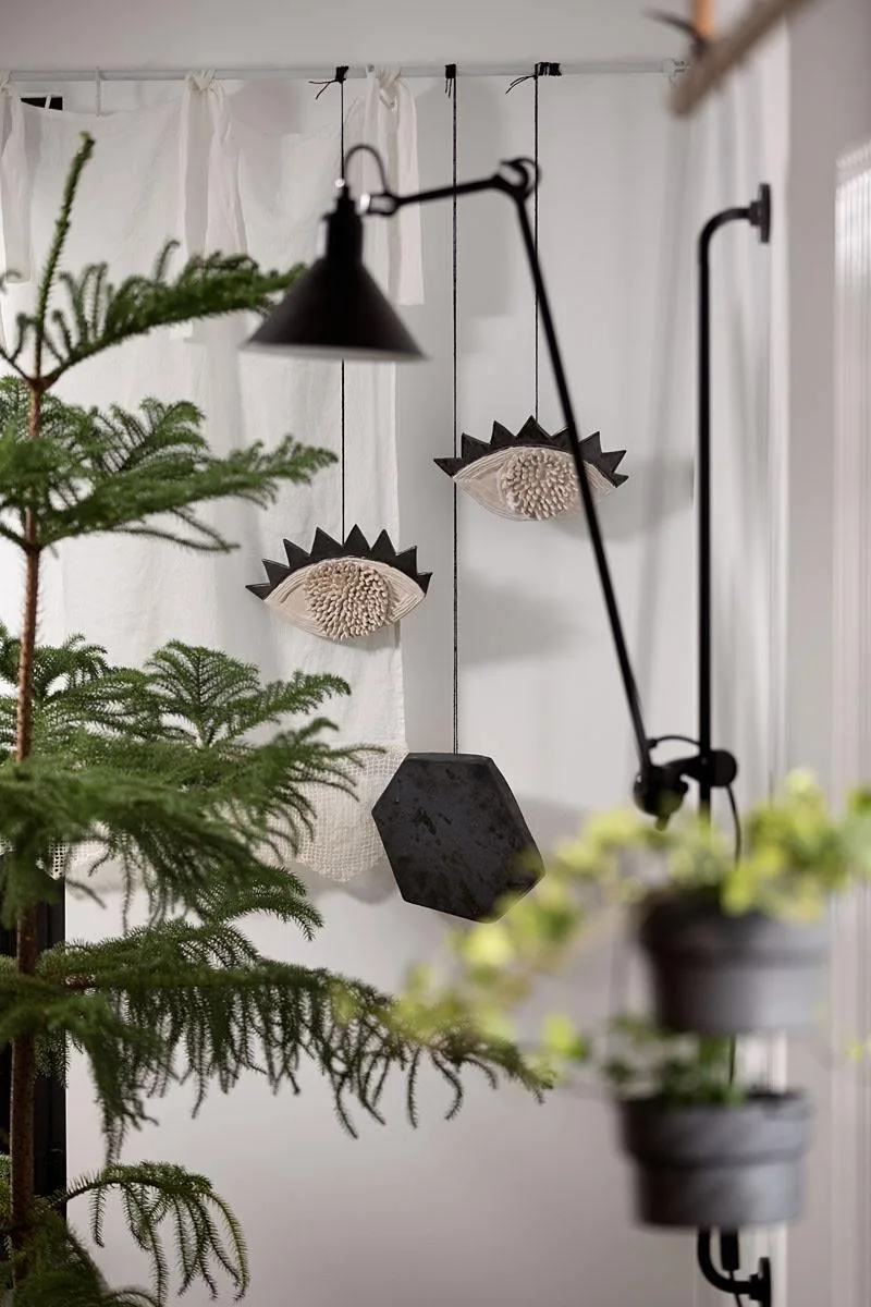 Two hanging eye-shaped sculptures and a black hexagon suspend from a white wall next to an adjustable black sconce and green plants.