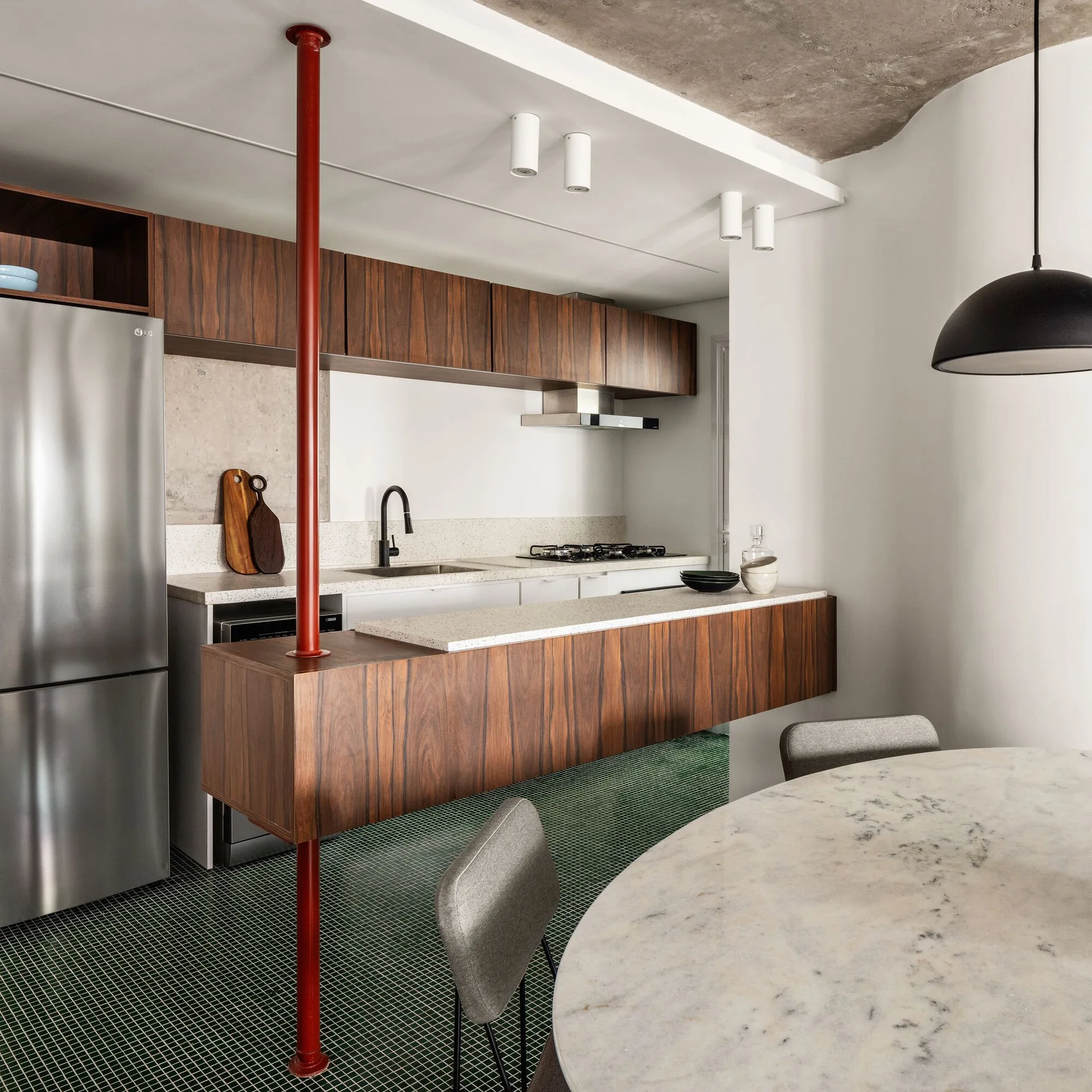 A modern kitchen with dark wood cabinets, a green mosaic tile floor, a red vertical support post, and a round marble dining table.