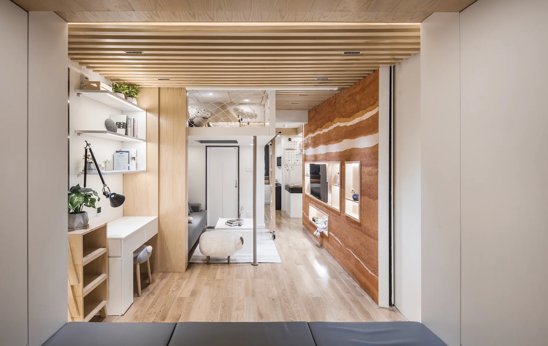 A small modern studio apartment showing a wooden slat ceiling, a layered terracotta earth feature wall, a built-in white desk, and a mezzanine loft.