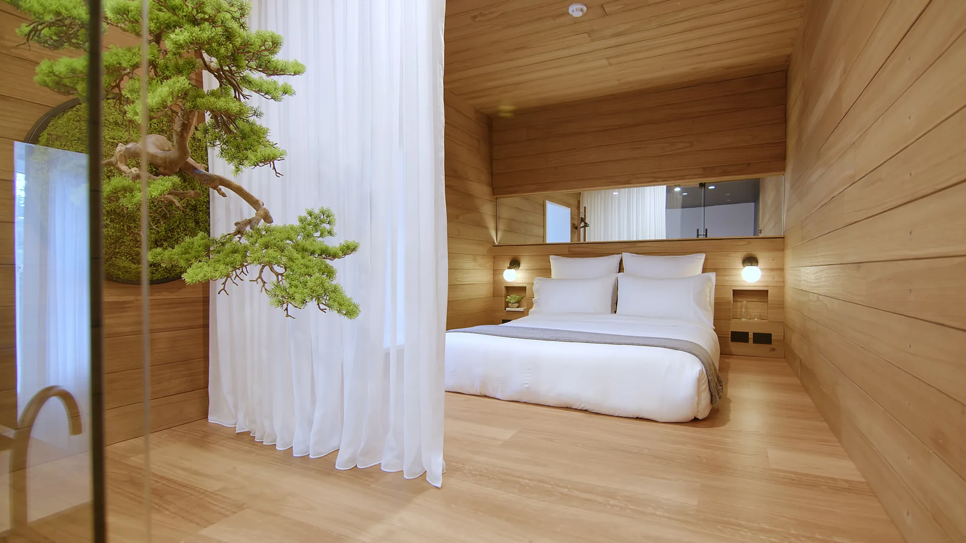 A wood-paneled bedroom features a bed with white linens, built-in sconces, a sheer white curtain divider, and a decorative bonsai tree.