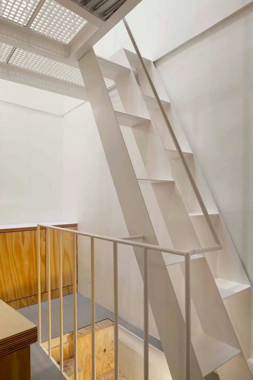 A minimalist white metal loft staircase rises beside slim white railings and natural plywood cabinets.