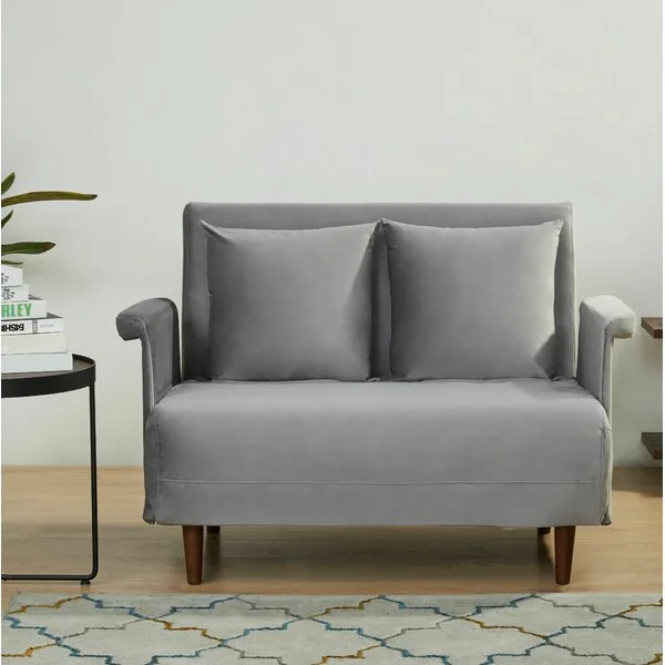 A grey fabric sofa bed with matching pillows and tapered wood legs stands on a light wood floor next to a small side table and patterned rug.