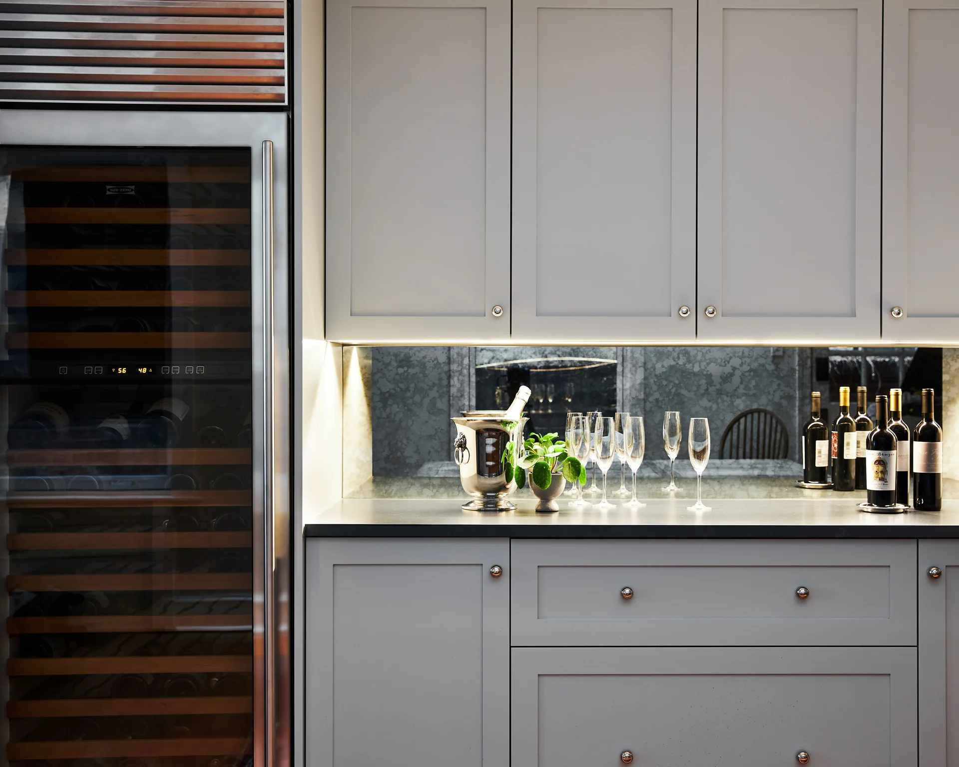 A grey home bar with shaker cabinets, an antiqued mirrored backsplash, under-cabinet lighting, and an integrated glass-front wine refrigerator.