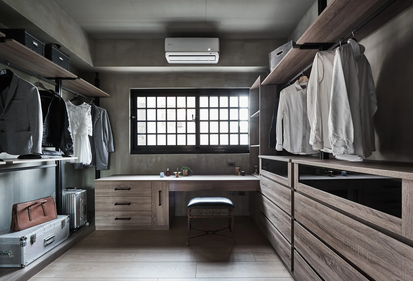 A walk-in closet with industrial concrete walls, wood-grained wardrobe storage, a built-in vanity desk under a grid window, and a leather stool.
