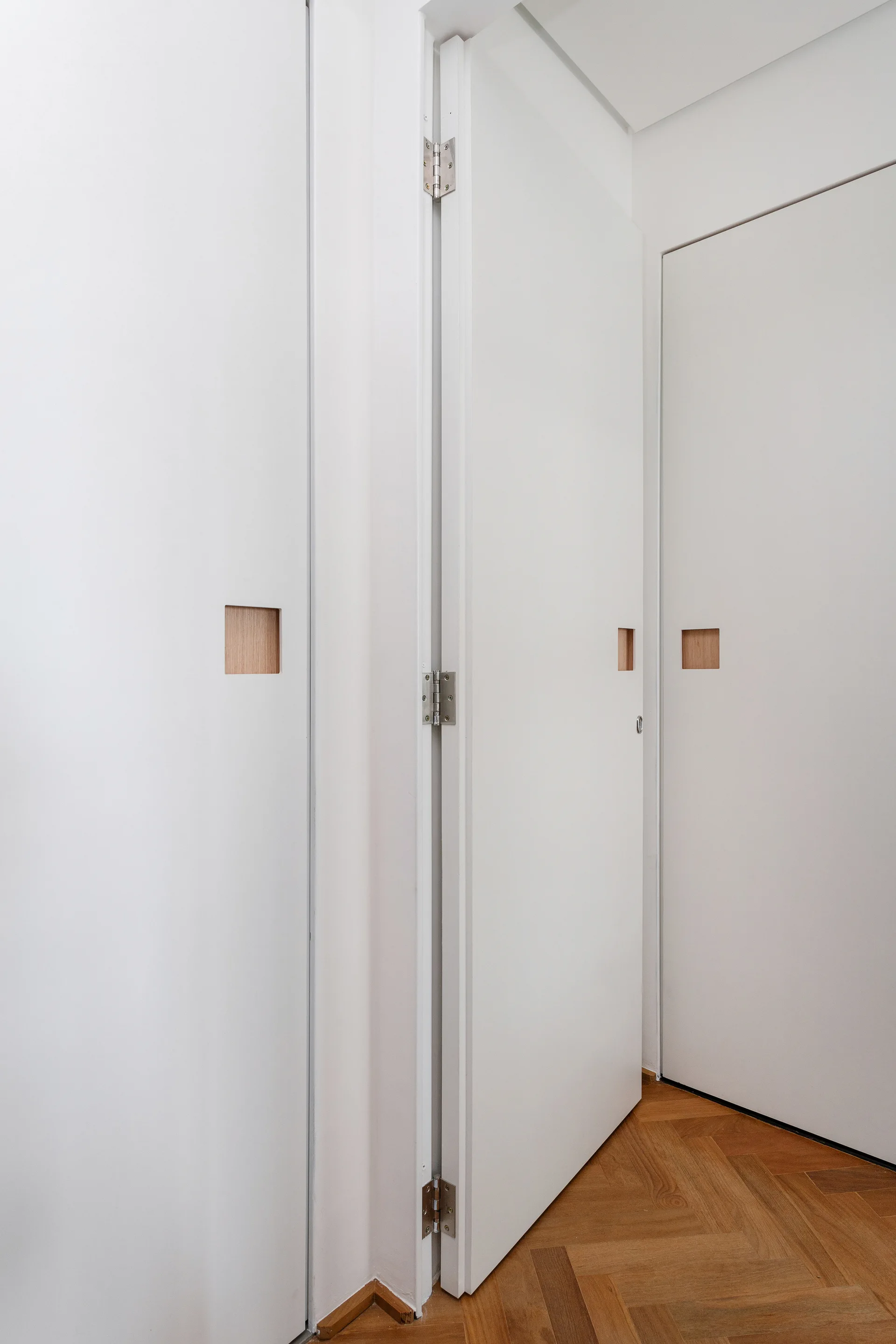 White minimalist doors featuring square wood-lined cutout handles are set above a warm herringbone parquet floor in a bright hallway.