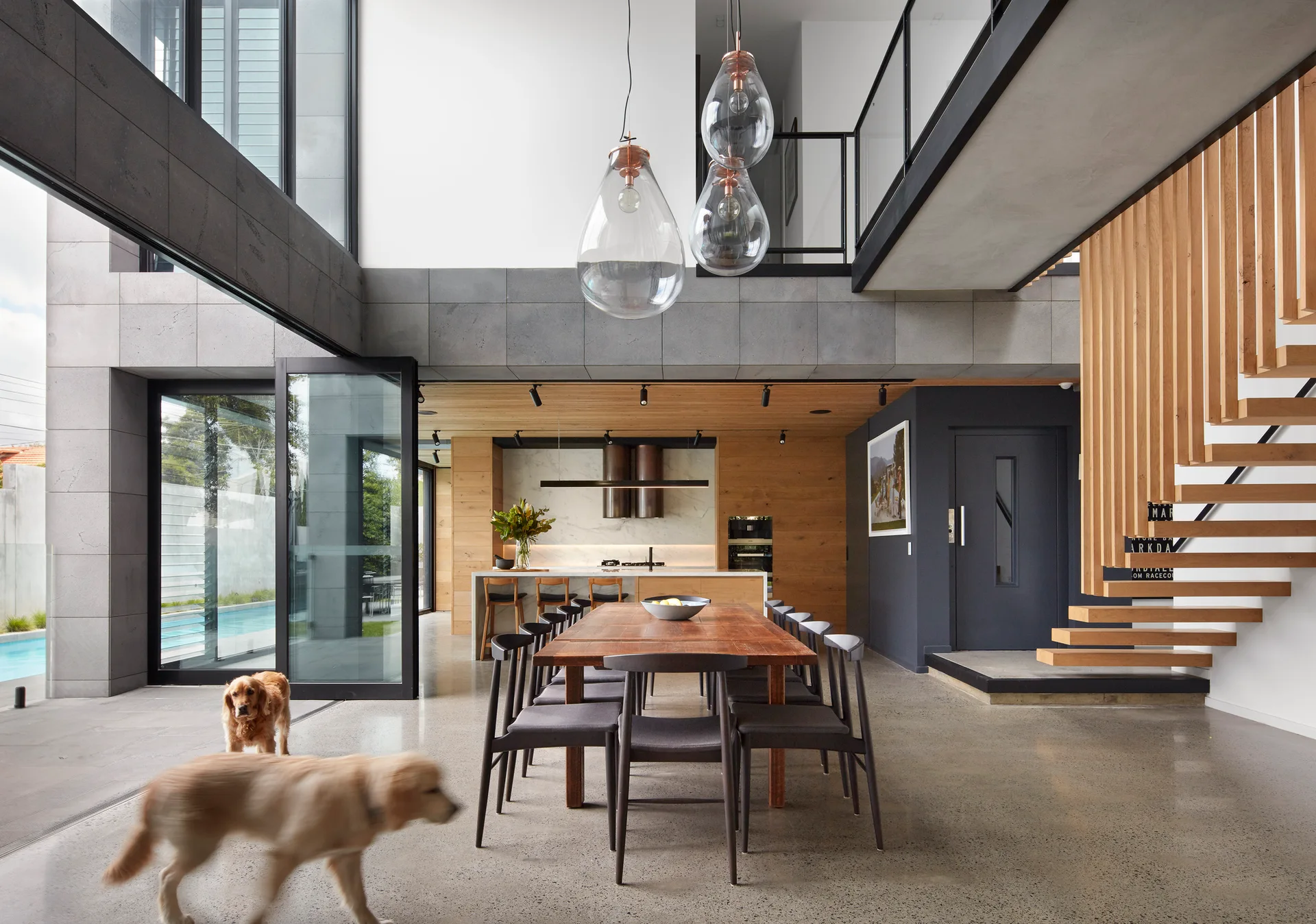 A modern open-plan dining room features a wooden table, a floating wood staircase, and oversized teardrop pendant lights below a high ceiling.