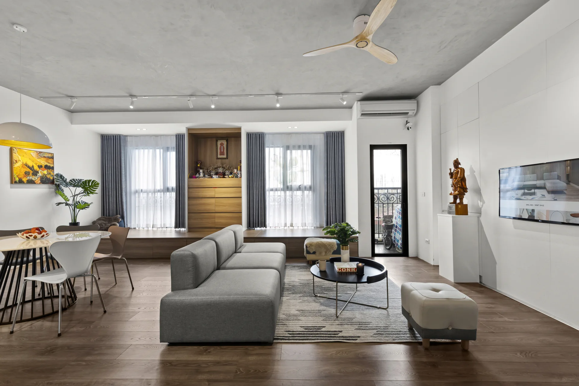 A modern open-plan living room featuring a grey sofa, a concrete ceiling, and low wooden built-in cabinets under two windows.