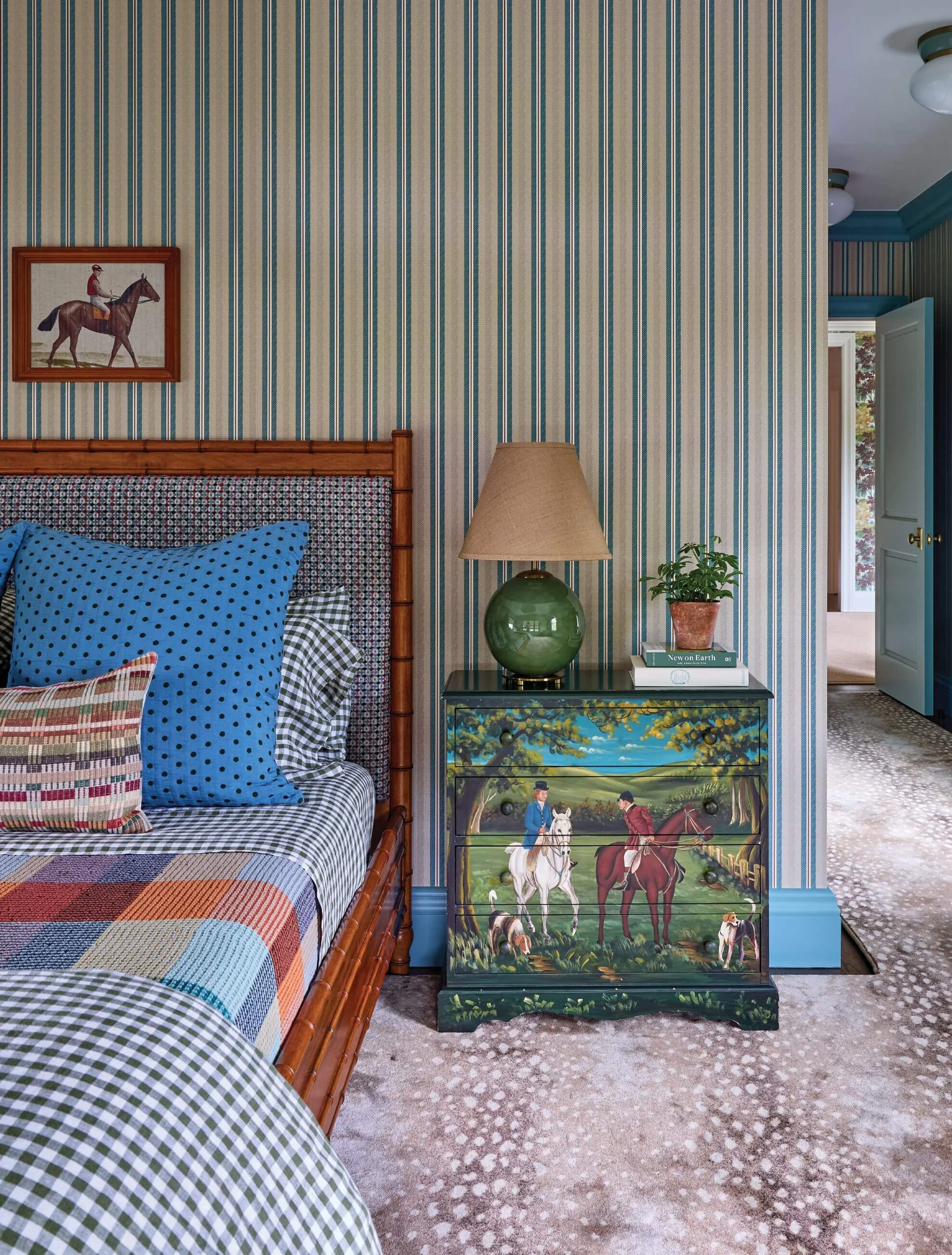 A maximalist bedroom features a faux bamboo bed with patterned linens next to a hand-painted equestrian chest against blue and green striped wallpaper.
