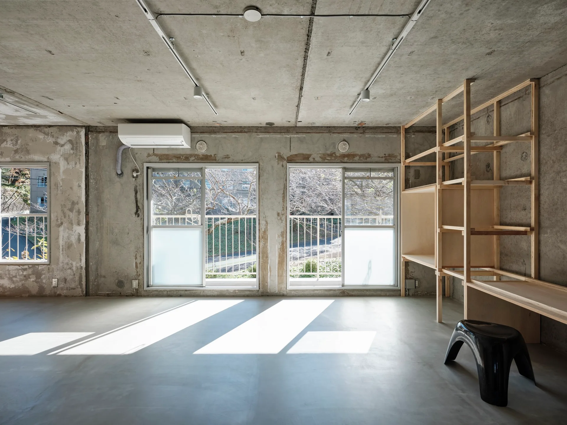 An industrial room features raw concrete walls and ceiling, a smooth concrete floor, large windows, and a tall open wood shelving unit.