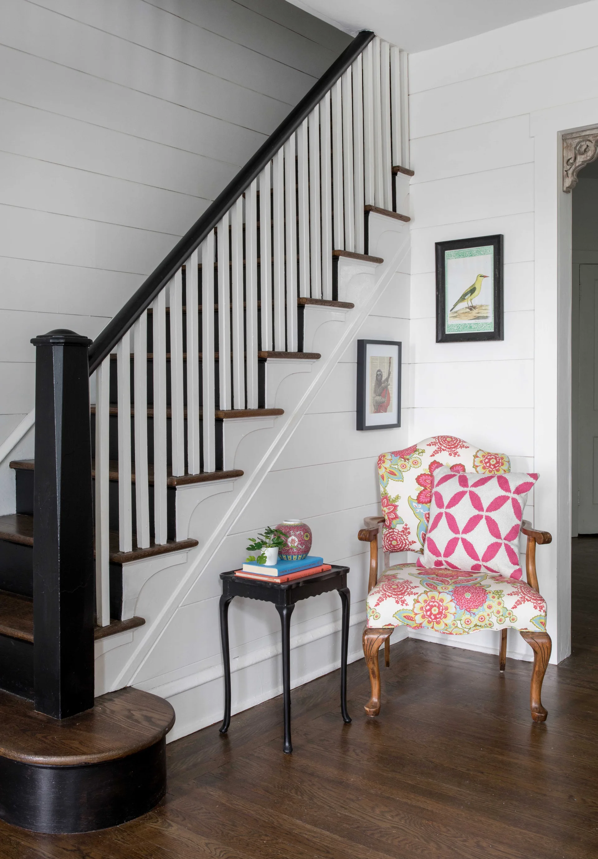 A wooden staircase with white shiplap walls and a black railing stands next to a small nook containing a floral armchair and a black side table.