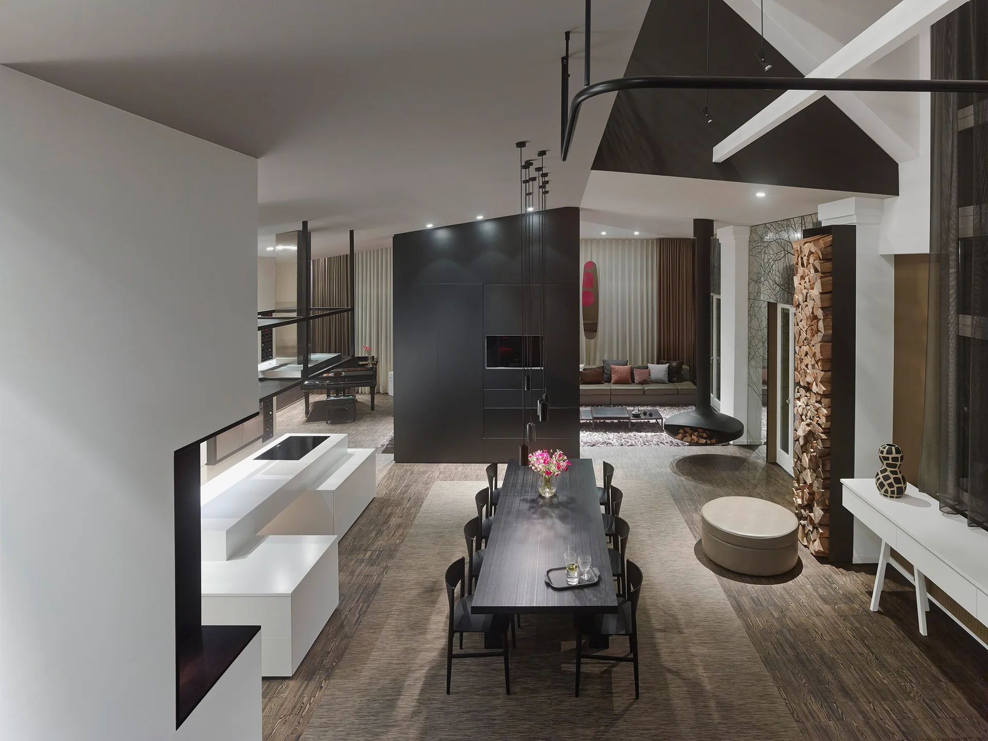High-angle view of an open-plan dining area featuring a long black table, a suspended black fireplace, and a tall vertical stack of logs.