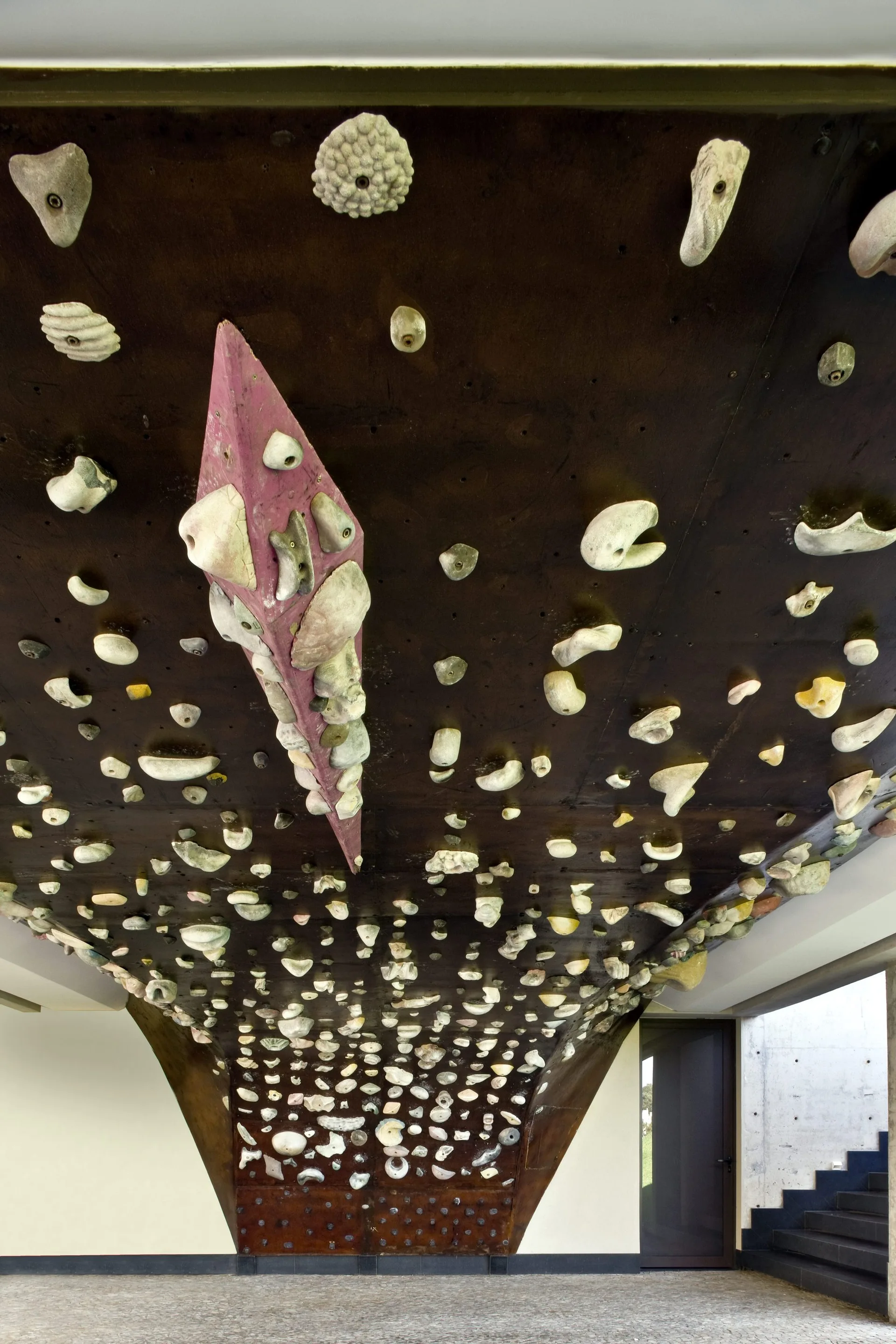 An indoor overhanging bouldering wall features dark wood panels and numerous textured climbing holds near a concrete staircase.