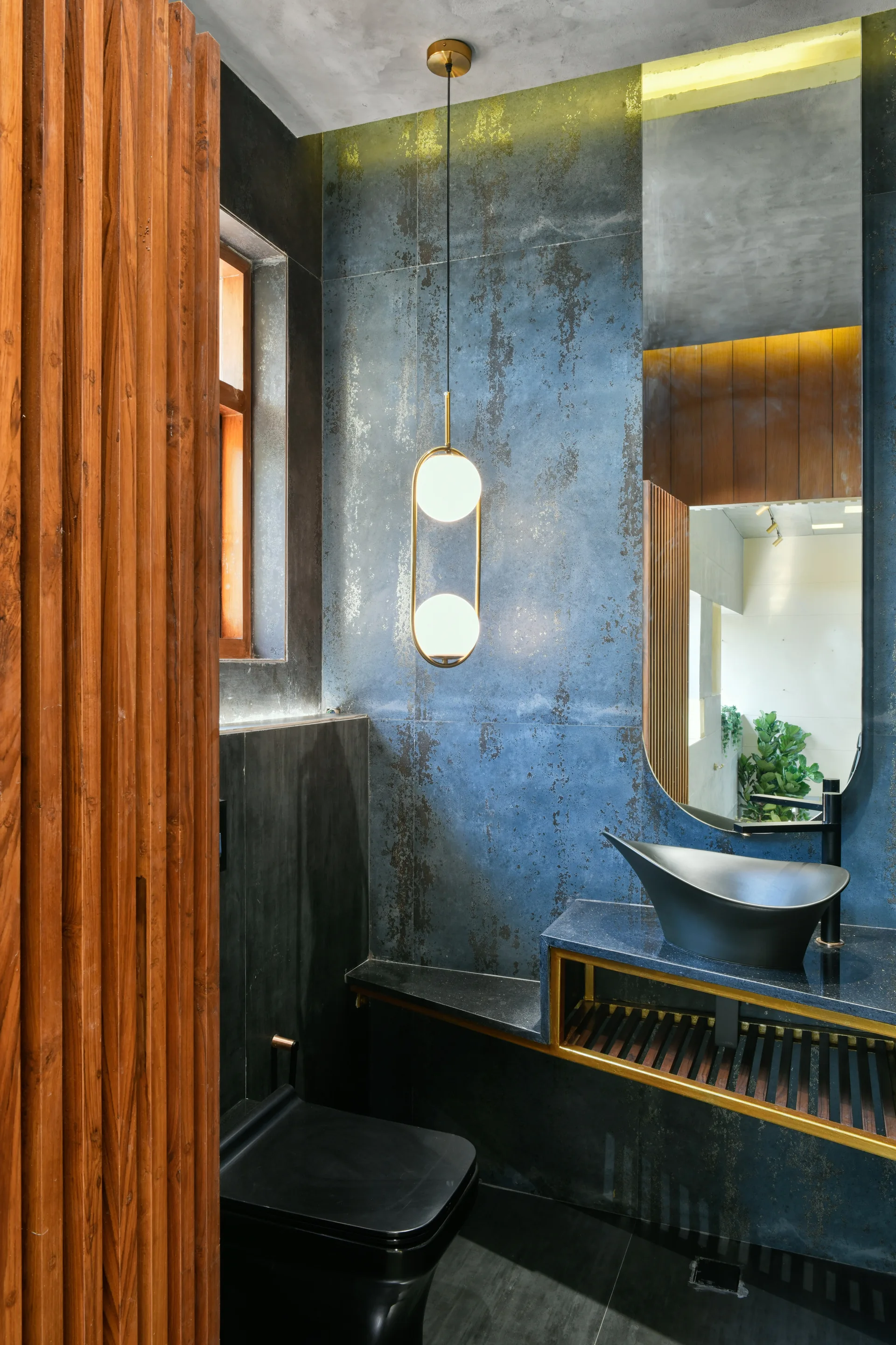 A modern bathroom features textured blue wall tiles, a black vessel sink on a dark stone vanity, a gold globe pendant light, and vertical wood slats.