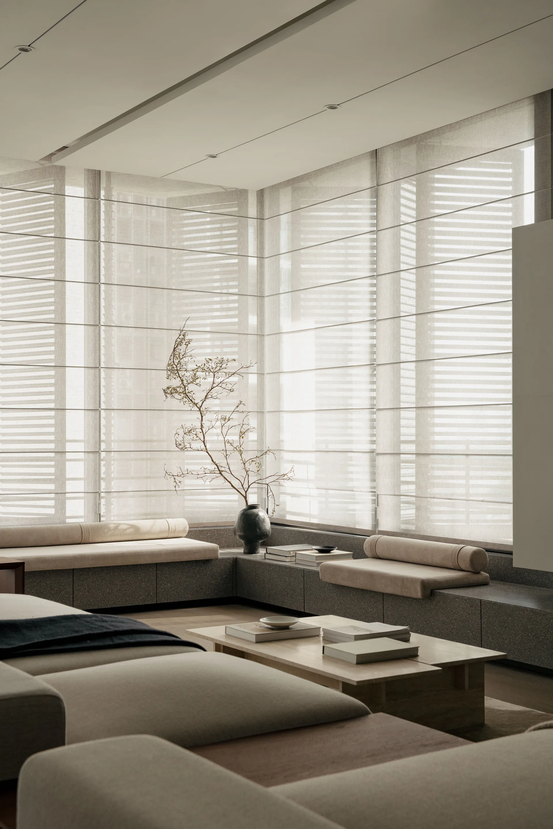A minimalist living room features a low stone-like corner banquette with beige cushions beneath windows fitted with translucent neutral Roman shades.