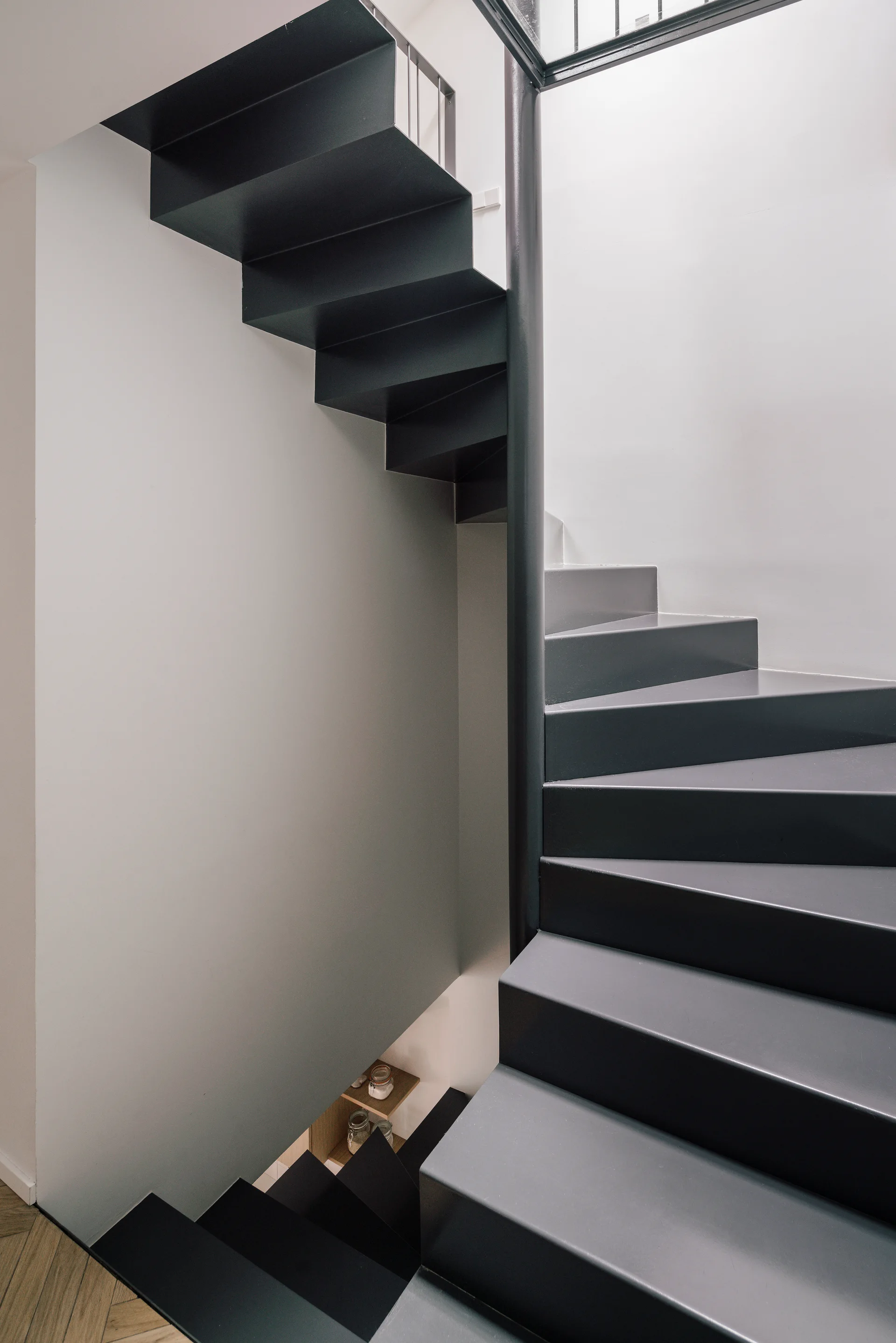 A dark gray geometric spiral staircase winds upward against smooth white walls, showing metal treads and a sliver of herringbone wood flooring.