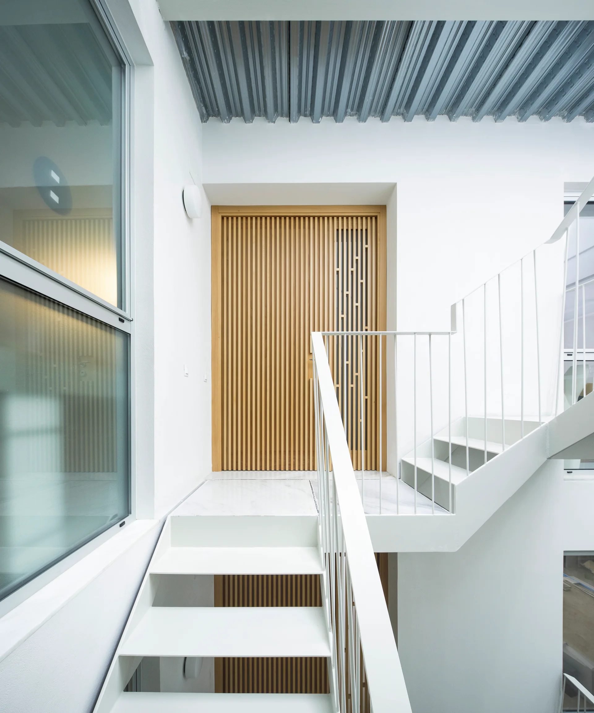 A white metal staircase leading to a landing with a veined floor and a light-toned slatted wood door under a corrugated metal ceiling.