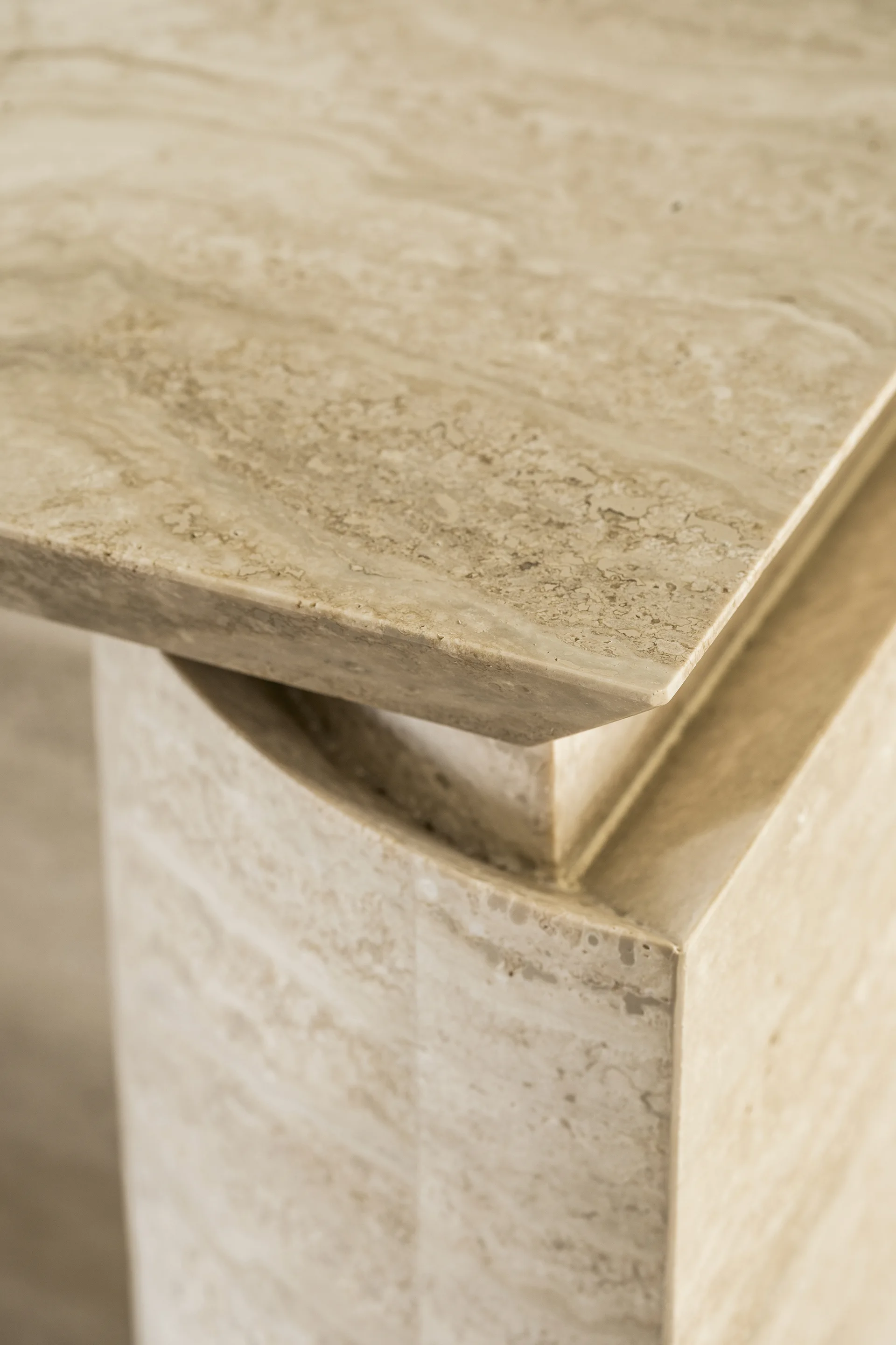 A close-up of a beige travertine table showing the matte textured top edge and a layered geometric pedestal base in natural light.