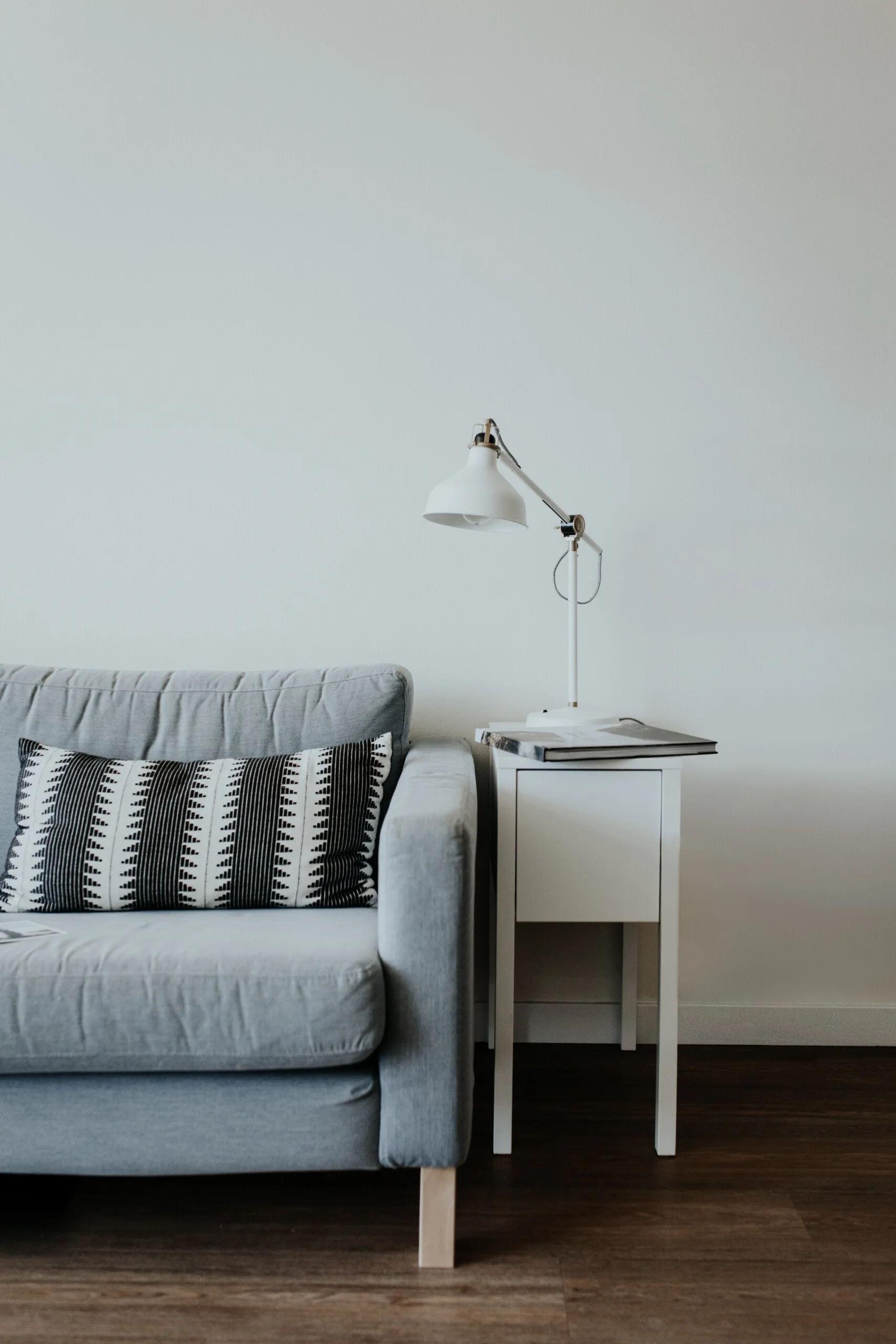 A grey fabric sofa with a black and white patterned pillow sits next to a white side table holding a white metal reading lamp on dark wood floors.