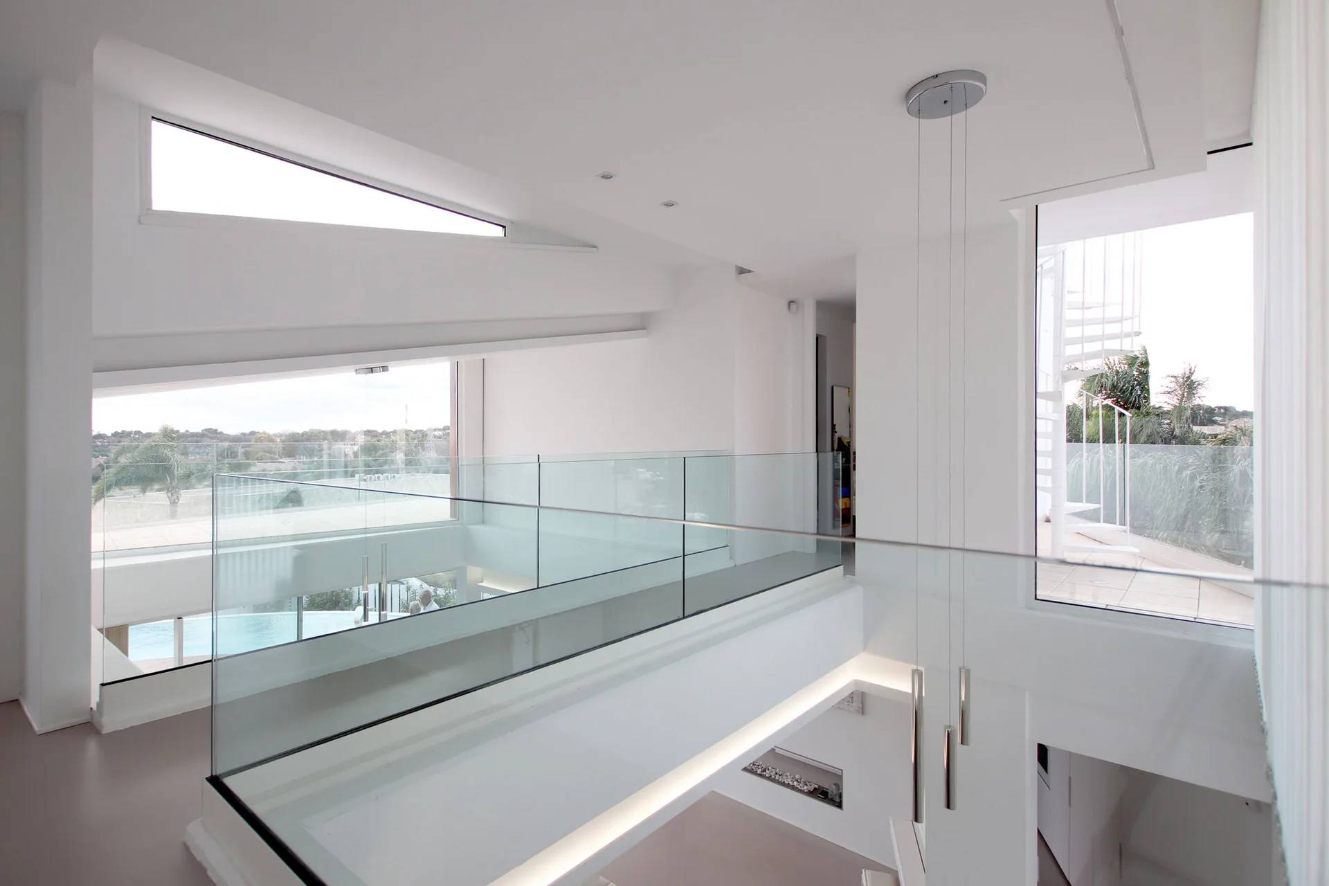 A white modern interior bridge features sleek glass railings, a sloped ceiling with an angular window, and slim cylindrical pendant lights.