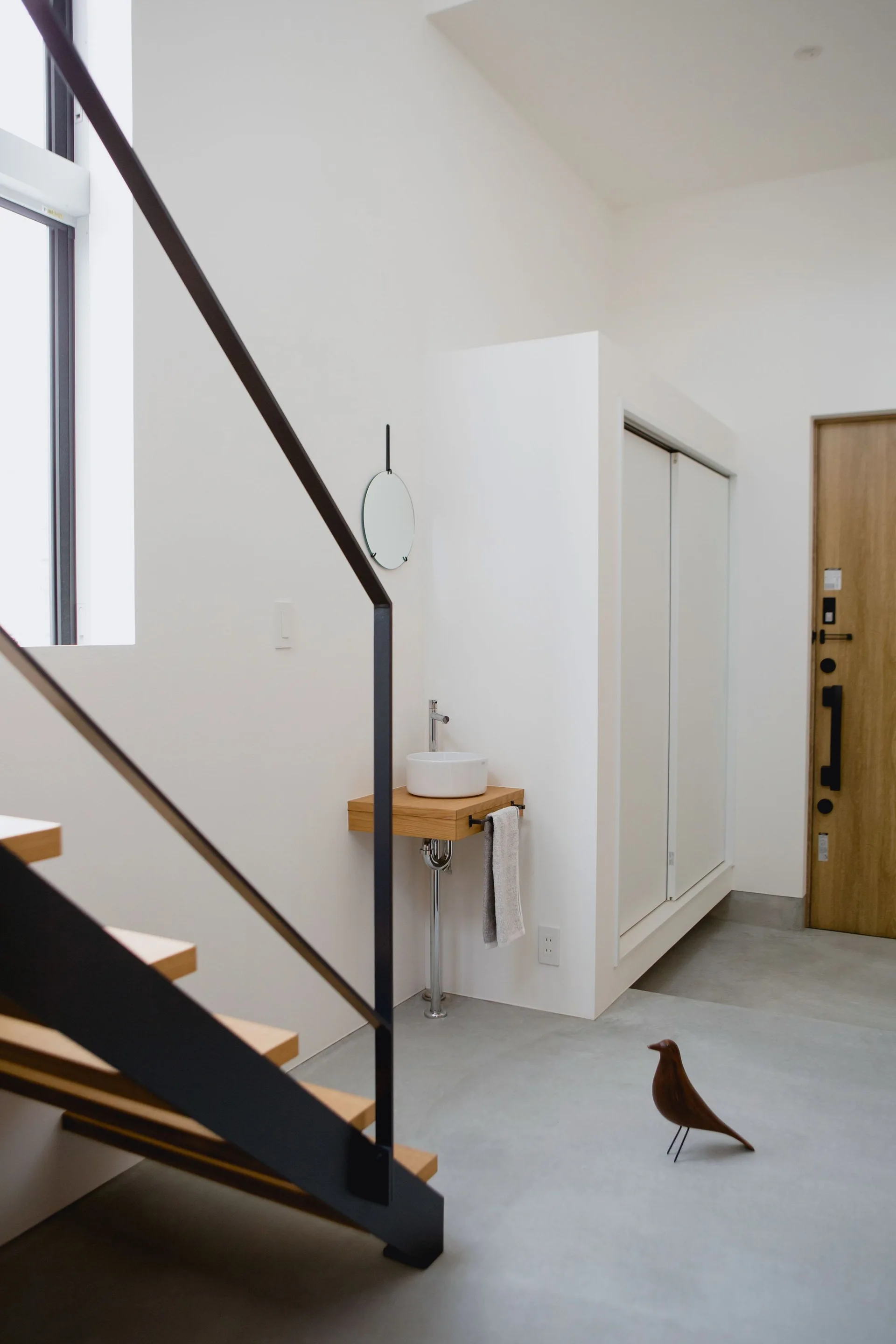 A minimalist entryway features a floating wood vanity with a ceramic vessel sink, a small round mirror, and a wood staircase.