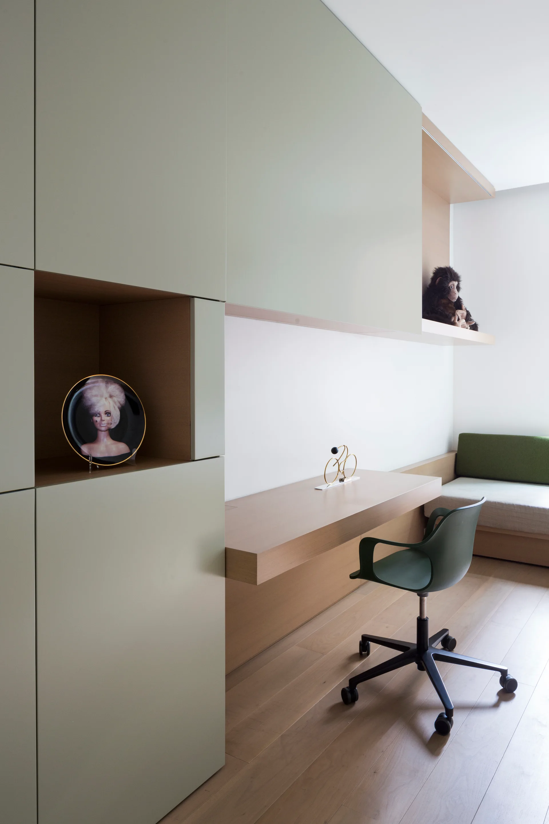 A contemporary study area with matte sage green storage cabinets, an integrated light wood desk, a matching green office chair, and light wood floors.
