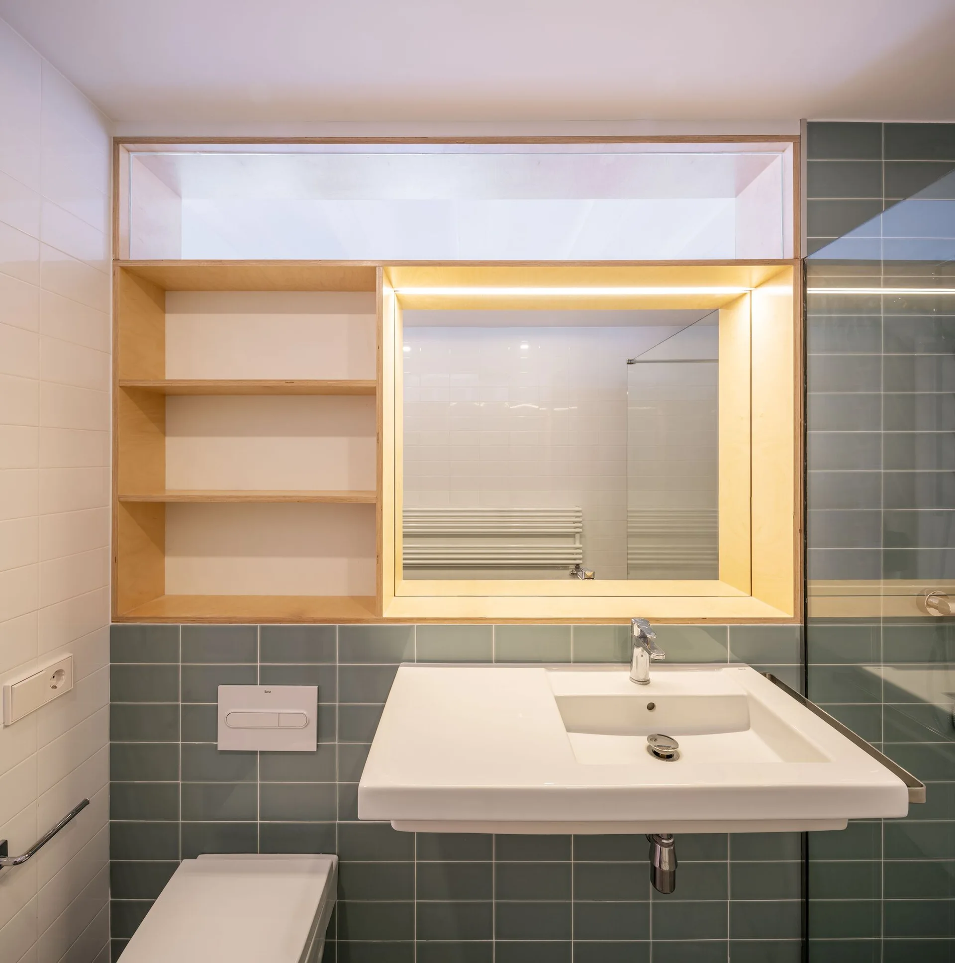 A modern bathroom features a white wall-mounted sink set against green square tiles beneath a light wood frame with open shelves and a backlit mirror.