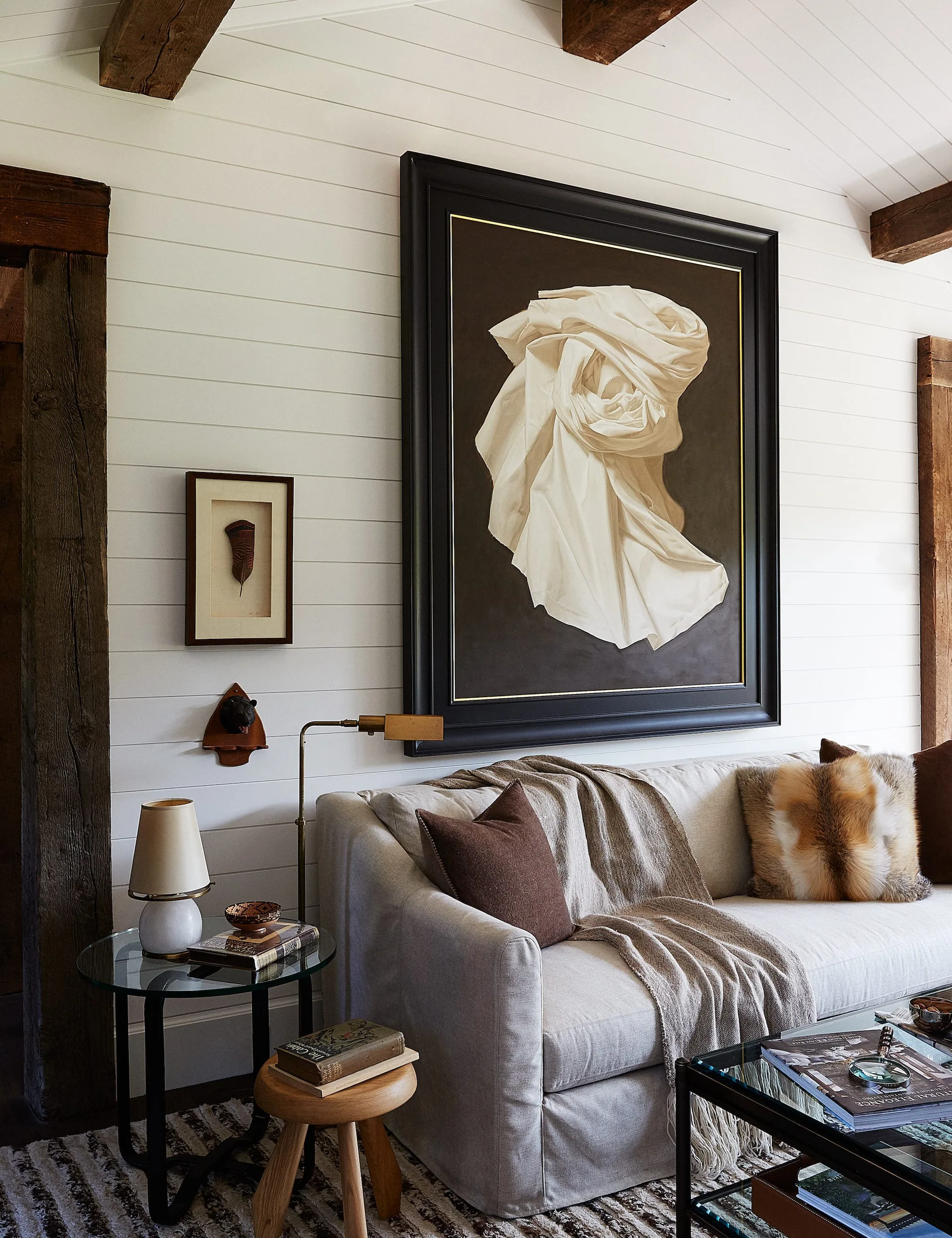 A grey sofa with brown and fur pillows sits against a white shiplap wall beneath a large framed painting of draped fabric and dark ceiling beams.