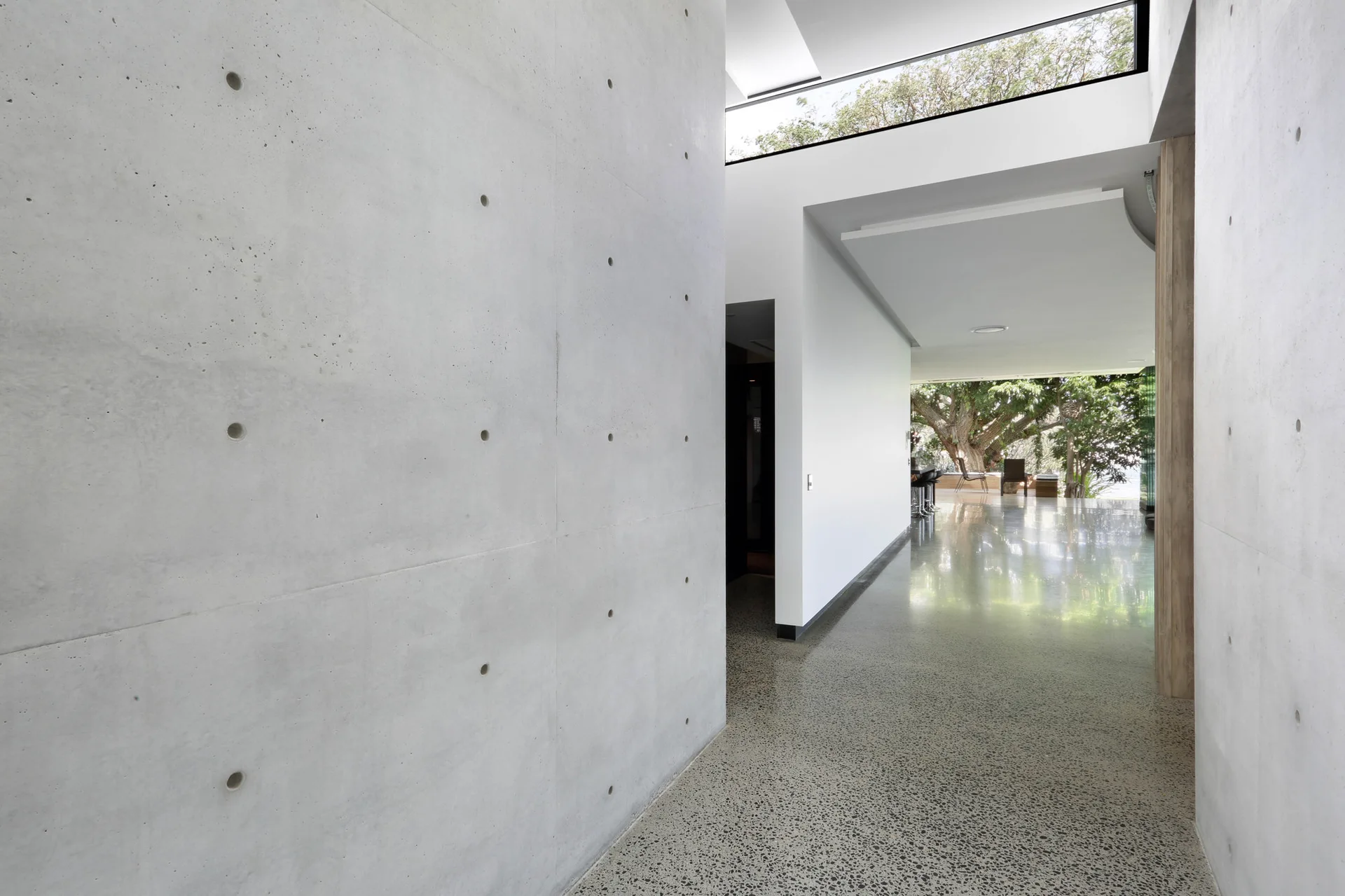 A minimalist residential hallway features high-gloss polished terrazzo flooring, exposed concrete walls with circular tie patterns, and a clerestory window.