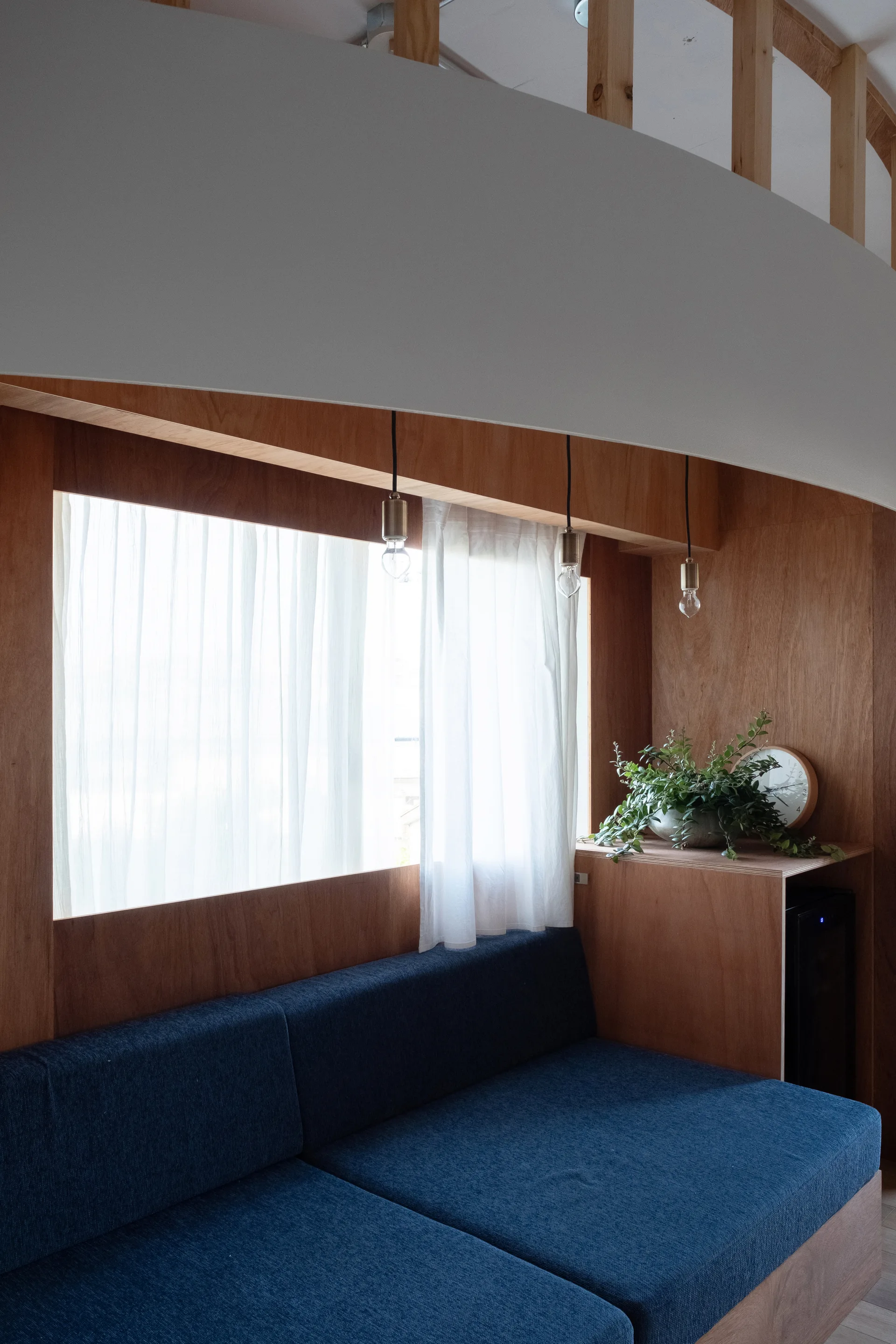 A built-in blue upholstered sofa sits in a warm plywood paneled nook with simple pendant lights hanging in front of a window.