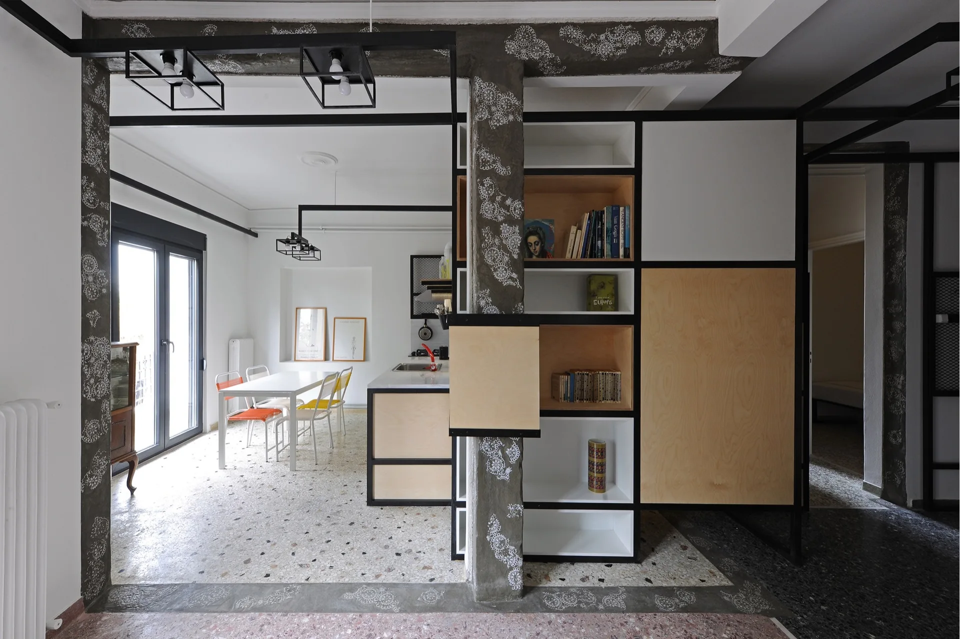A modern open-concept dining area and kitchen separated by a black steel and plywood shelving unit on terrazzo flooring.
