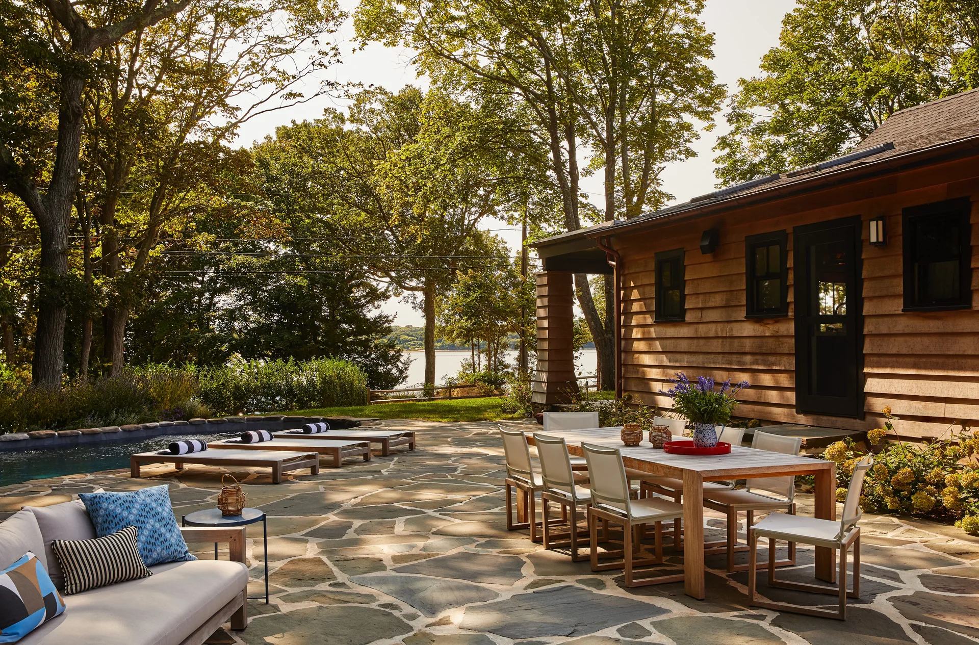 A spacious flagstone patio features a wooden dining table, a lounge sofa with blue pillows, and wooden pool chairs beside a swimming pool.