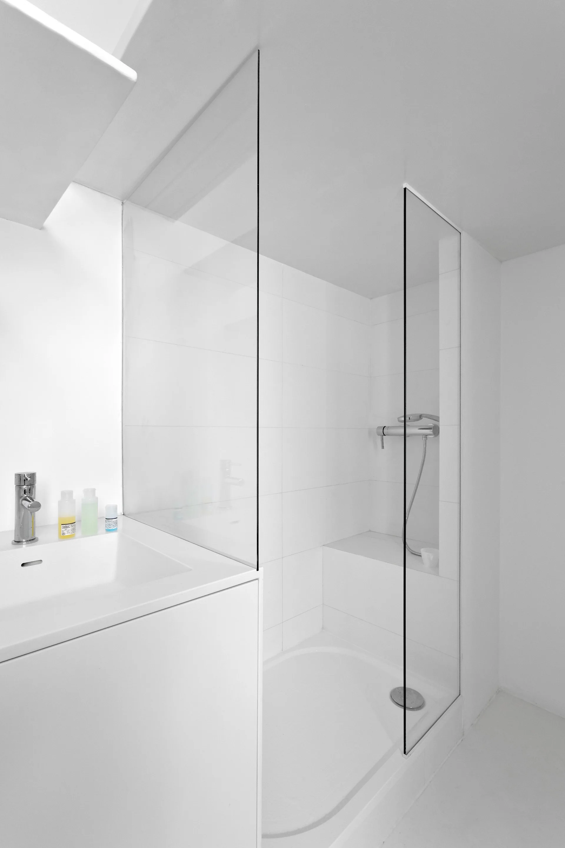 A minimalist white bathroom featuring a walk-in shower with a frameless glass partition, a built-in bench, and an integrated vanity sink.