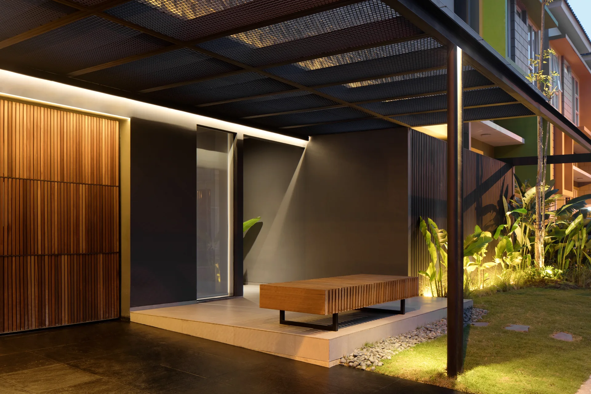 A modern outdoor porch features a wooden slatted bench, a black metal mesh canopy, and integrated warm lighting reflecting on dark grey walls.