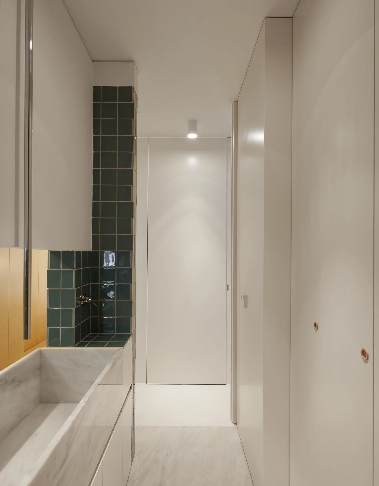 A narrow minimalist bathroom features a long white marble sink, a column of dark green square tiles, and seamless floor-to-ceiling white cabinetry.