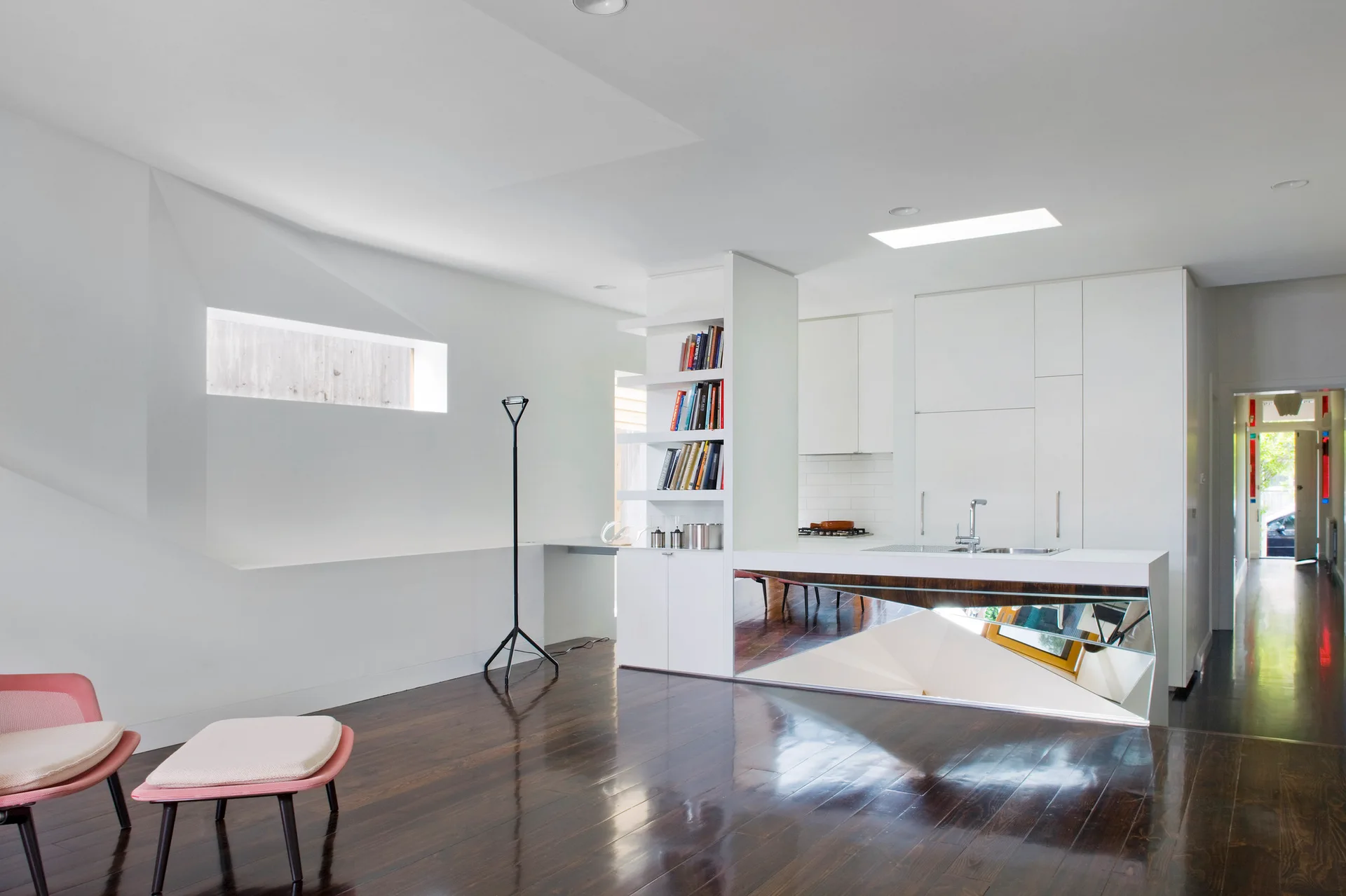 A minimalist white kitchen features a unique mirrored island base, dark wood floors, and a built-in vertical bookcase.