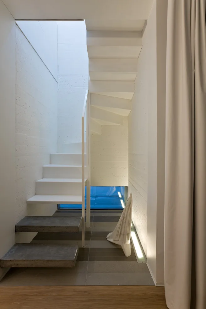 A minimalist white staircase ascends next to a textured concrete wall with a low window at the bottom.