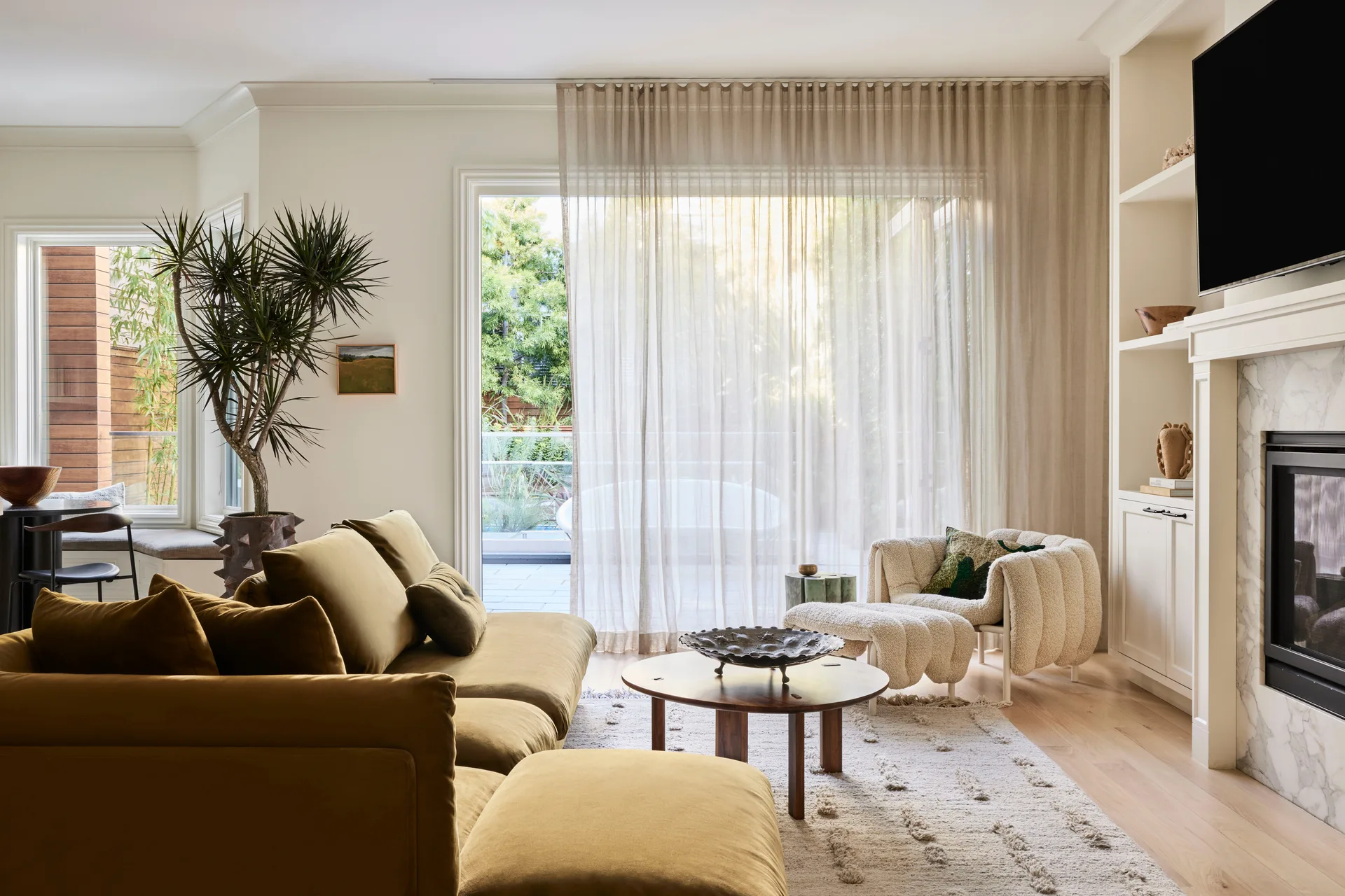 A light-filled living room features an olive green sectional sofa, a textured white armchair, and a marble fireplace integrated into white shelving.