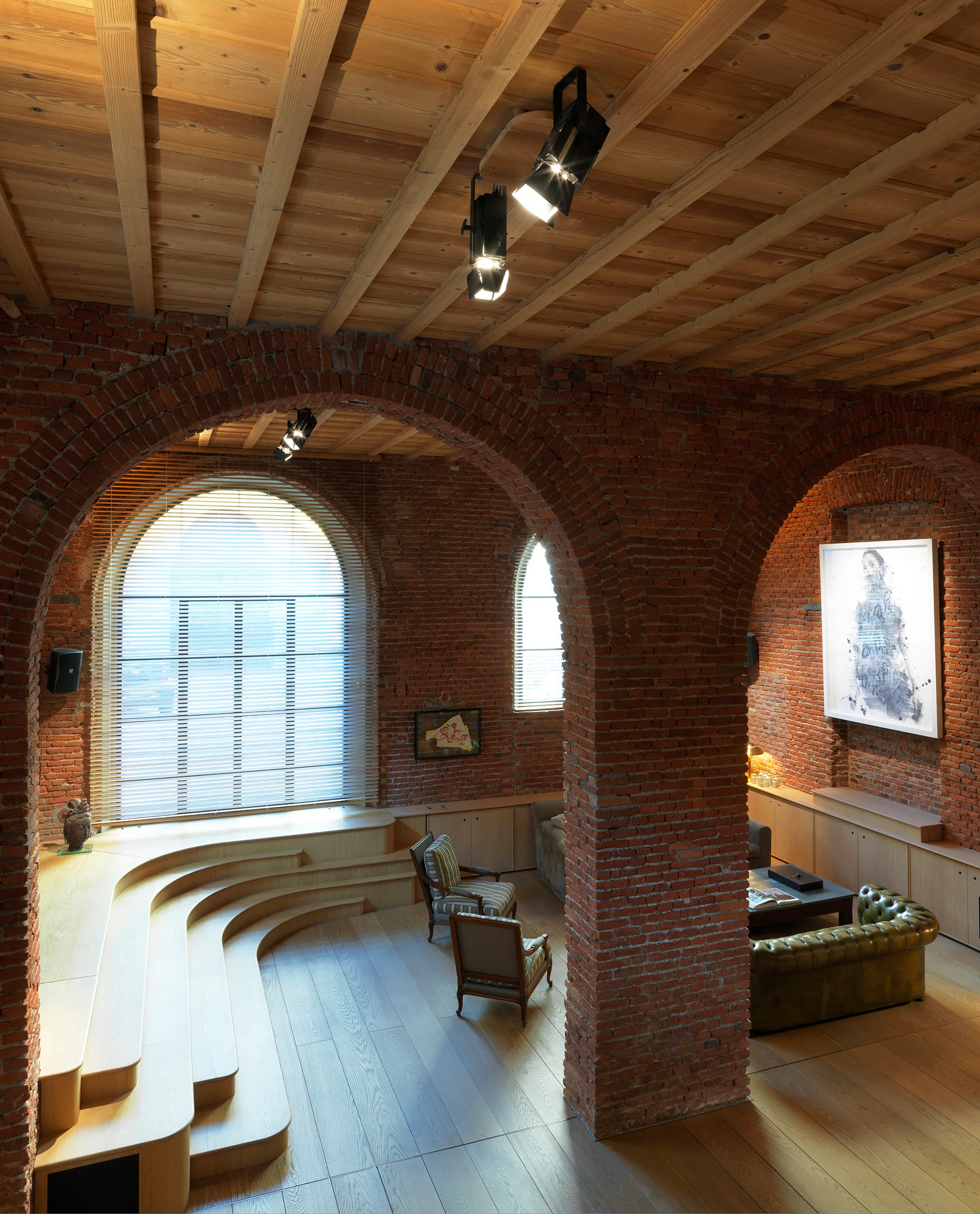 An industrial living space features structural red brick arches, a curved timber stepped platform, and a tufted green leather sofa.