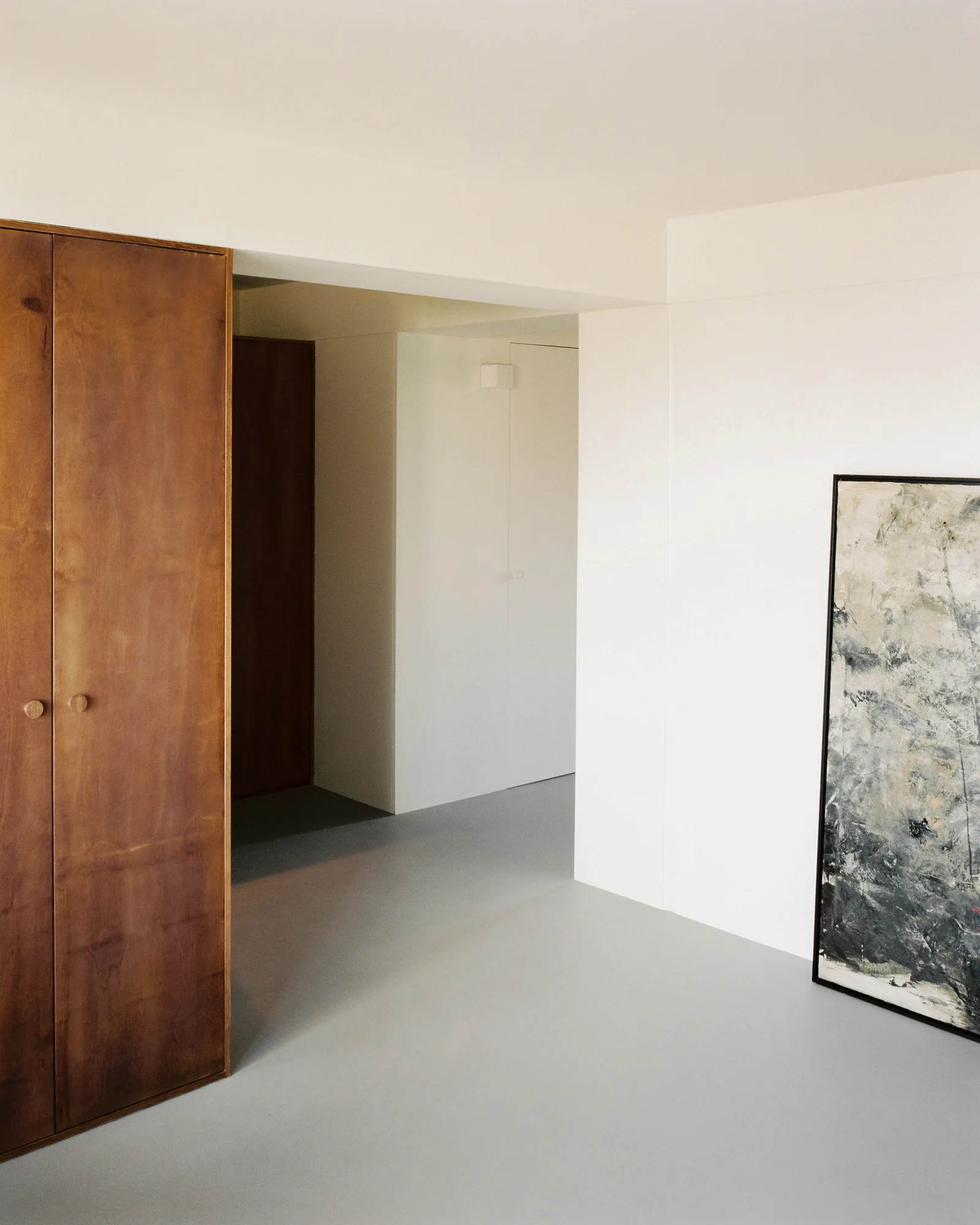 A minimalist hallway features tall warm wood cabinetry, matte white walls, seamless grey flooring, and a large abstract painting leaning against the wall.