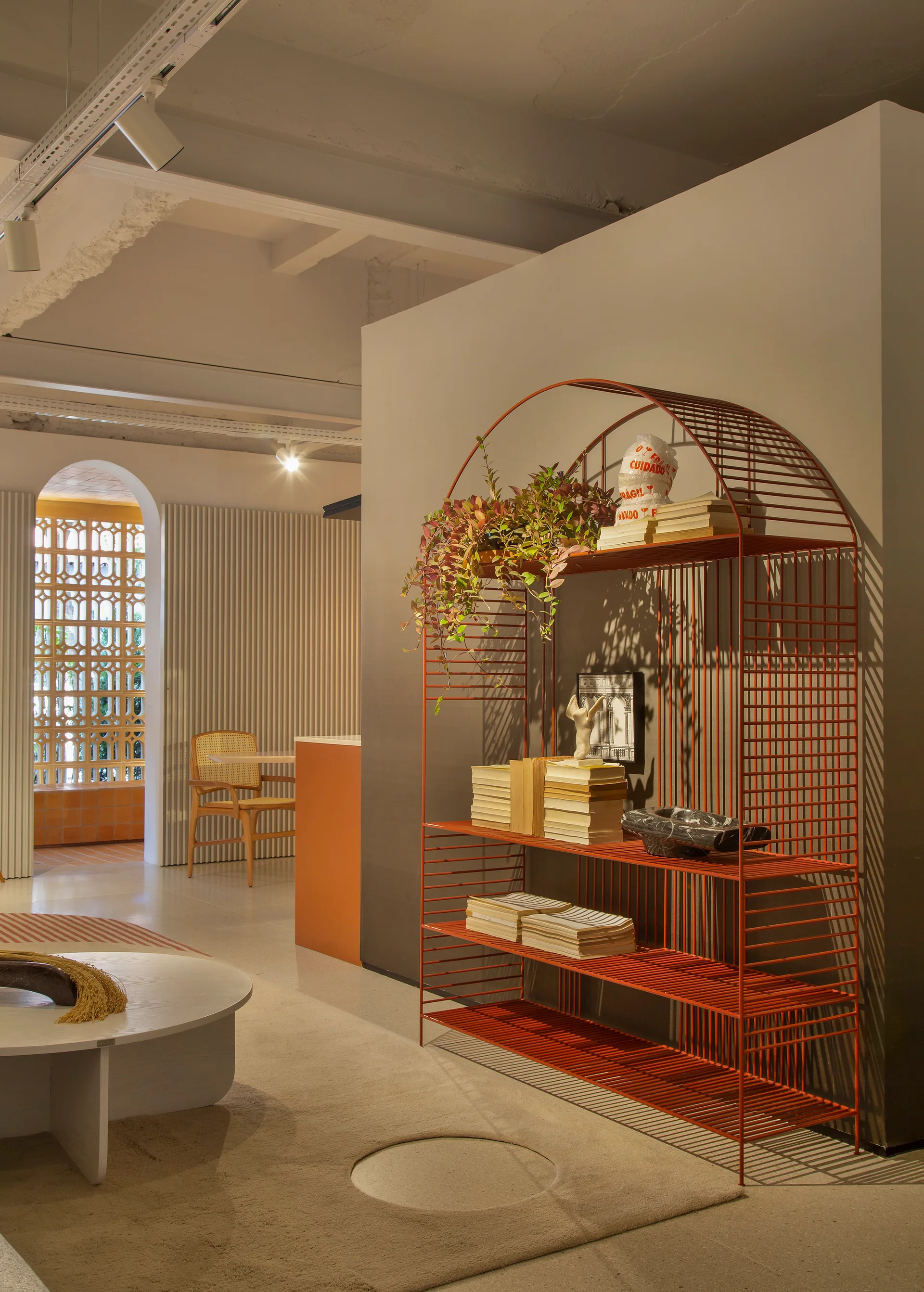 A tall terracotta wire shelving unit stands next to a low white coffee table in a modern interior with a slatted wall partition and background archway.
