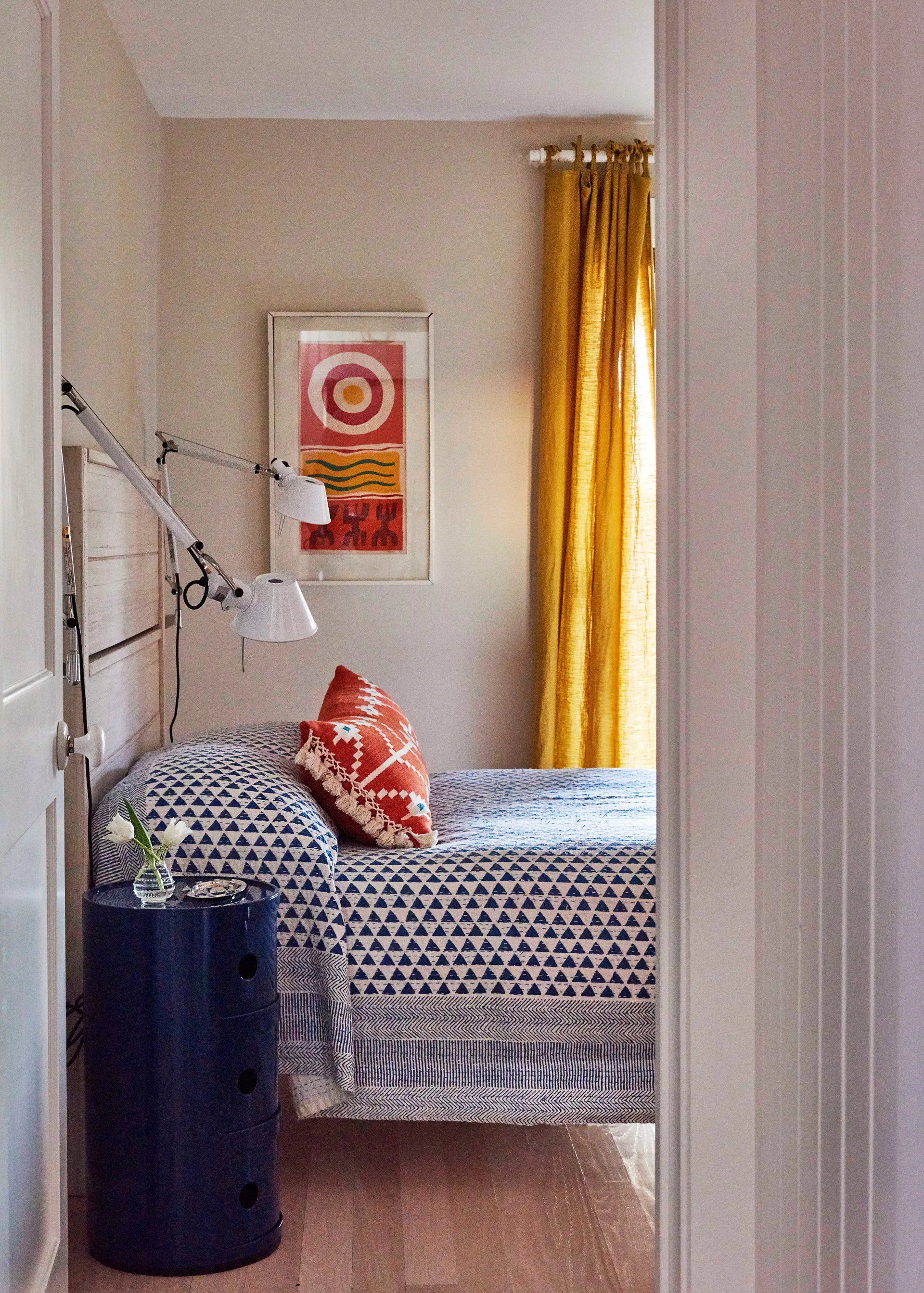 A small bedroom viewed through a doorway, featuring a bed with a blue patterned quilt, a glossy blue cylindrical nightstand, and yellow curtains.