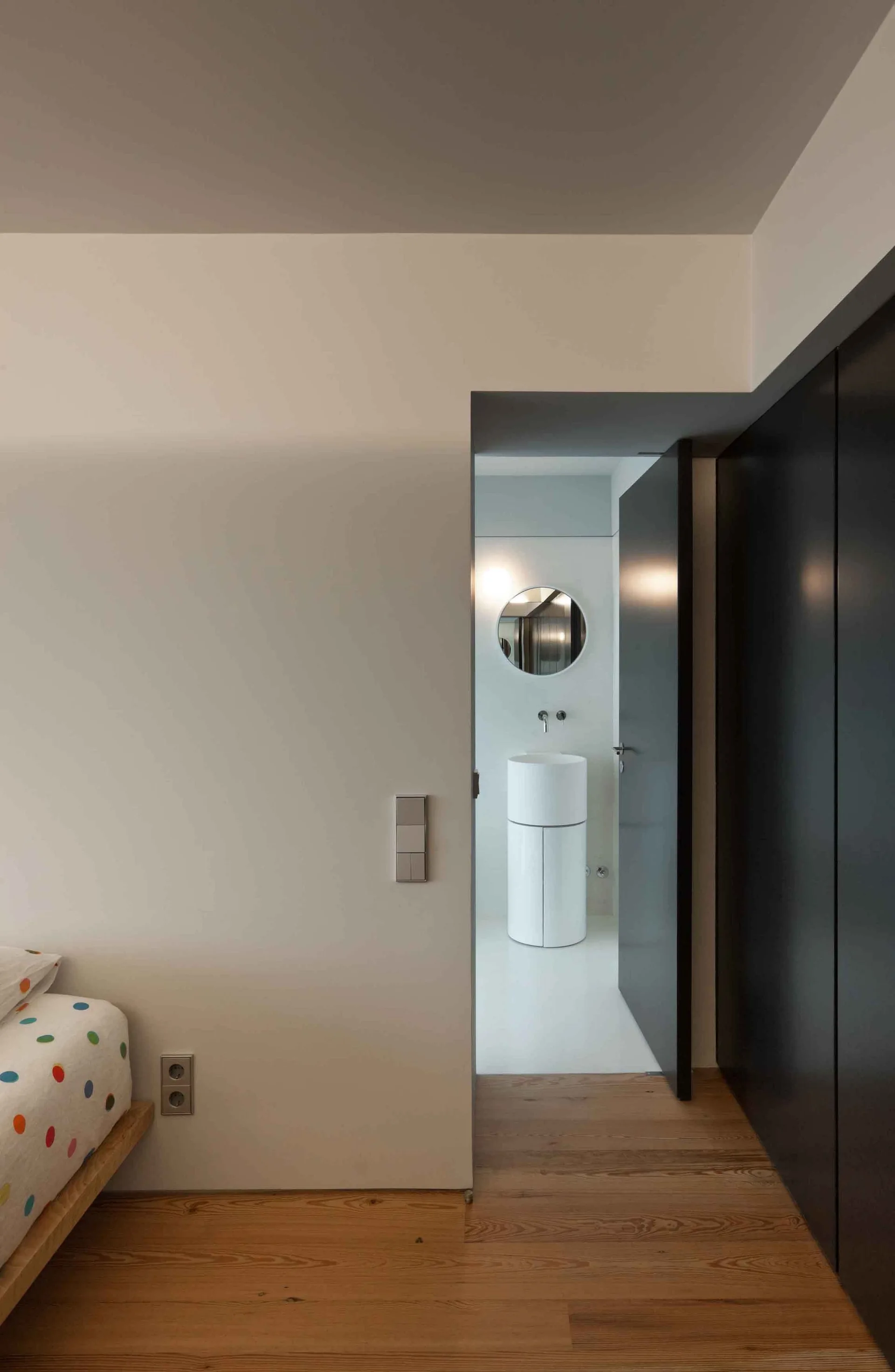 A view through a bedroom doorway into a minimalist bathroom featuring a white cylindrical pedestal sink and a round mirror.