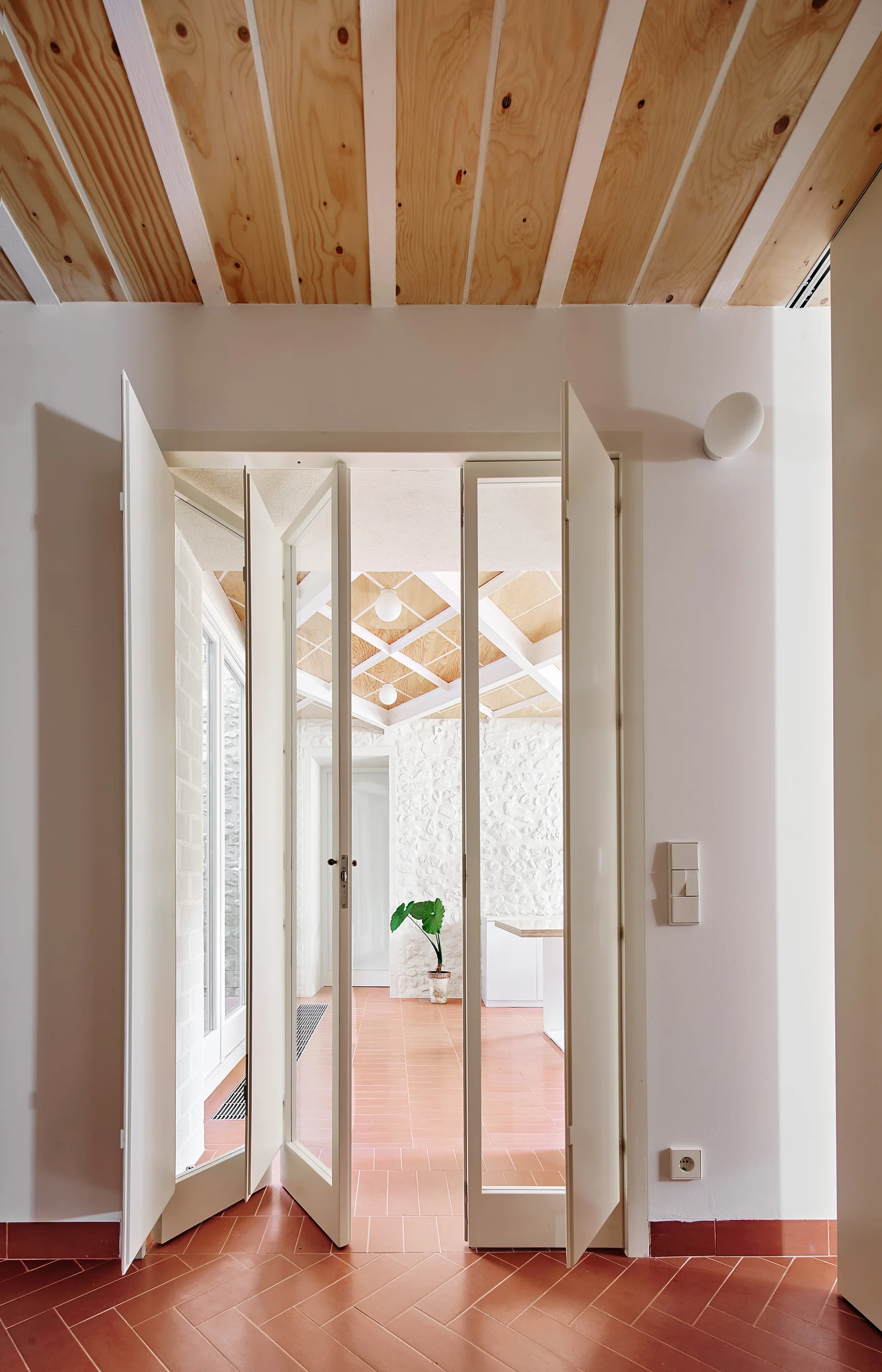 A view through open white folding glass doors from a herringbone terracotta tile floor into a room with stone walls and a wood-beamed ceiling.