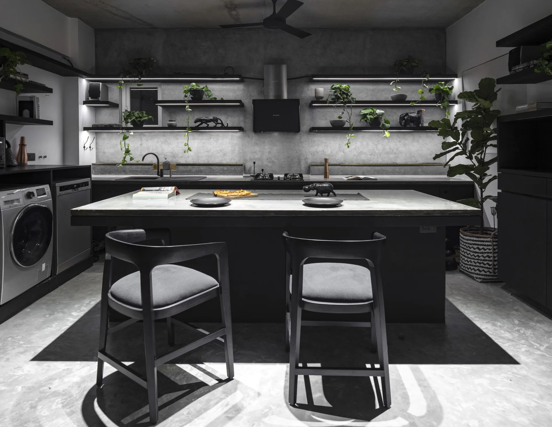 An industrial kitchen featuring concrete walls, black floating shelves with integrated LED lights, and a stone-topped island with two black stools.