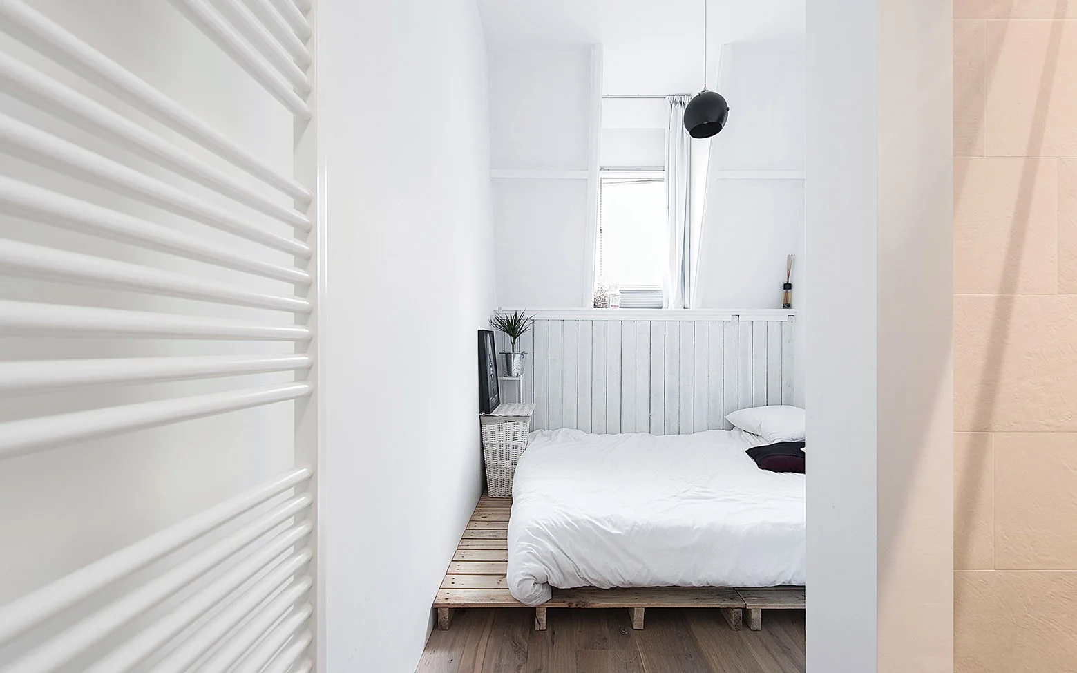 A narrow minimalist bedroom features a low wooden platform bed, white vertical wall paneling, a small window, and a black pendant light.