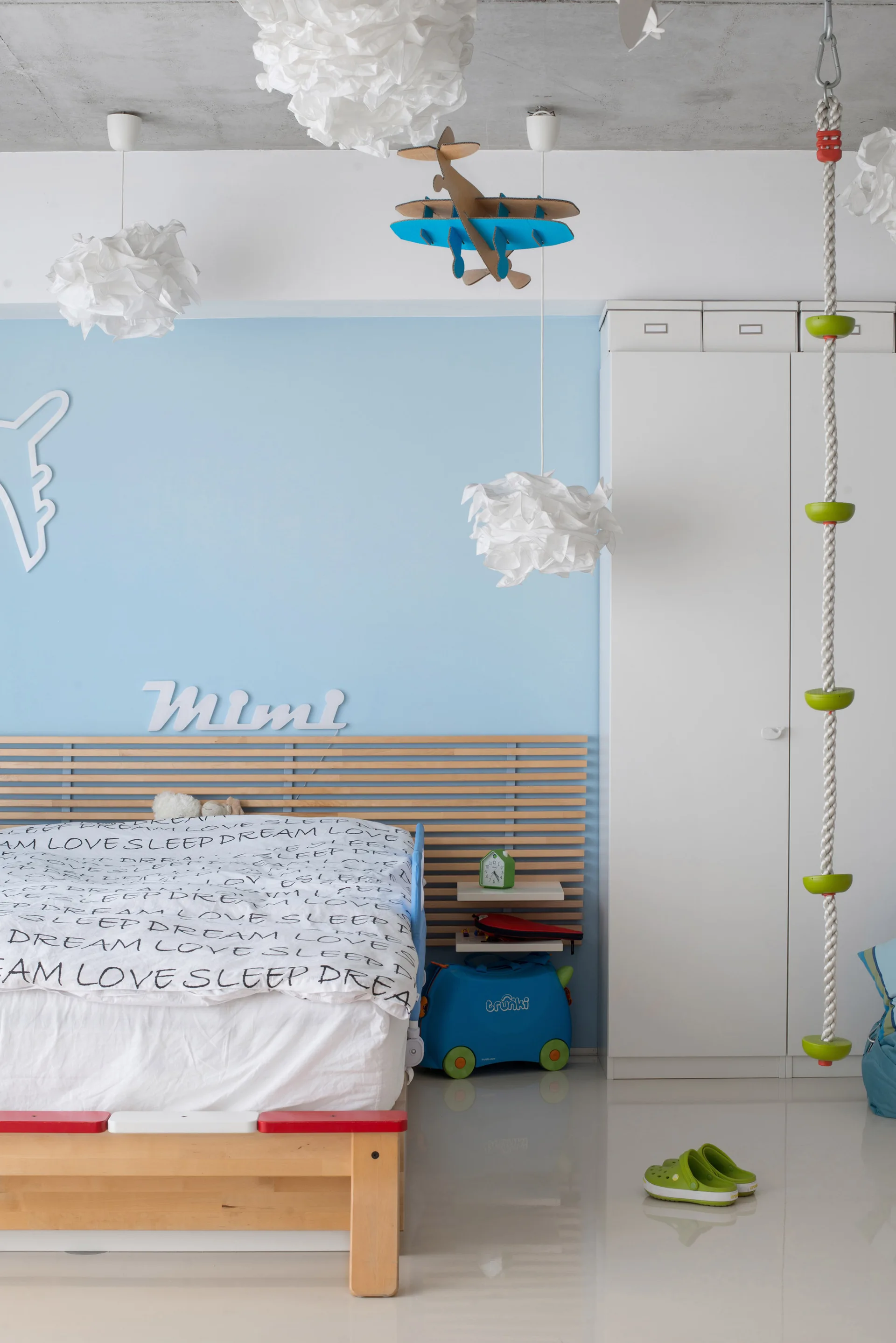 A kids bedroom features a bed with a wide wood slatted headboard, cloud-like pendant lights, and a climbing rope next to a wardrobe.